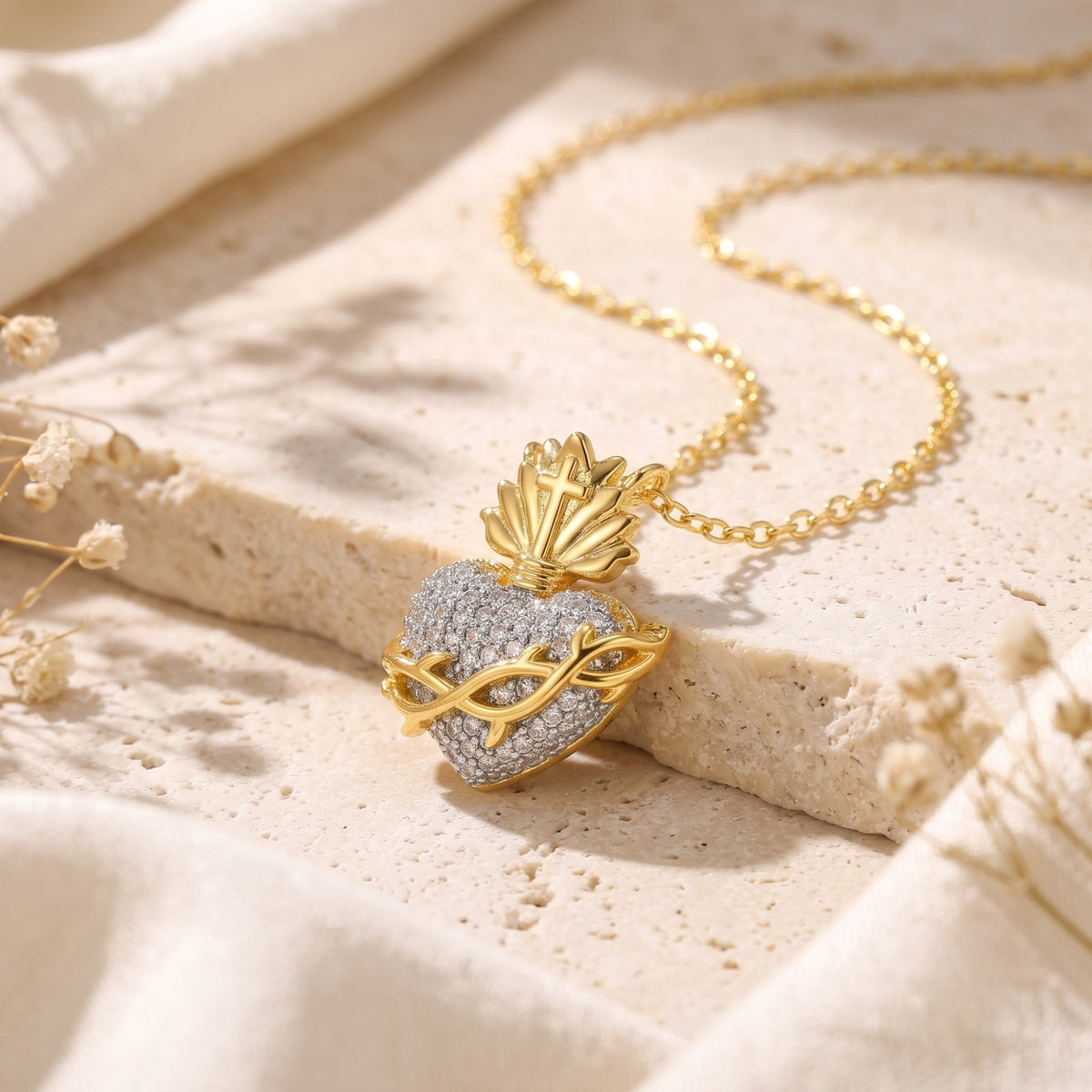 Angled gold-tone sacred heart pendant with a thorn-like vine and cubic zirconia, resting on a pale stone surface.