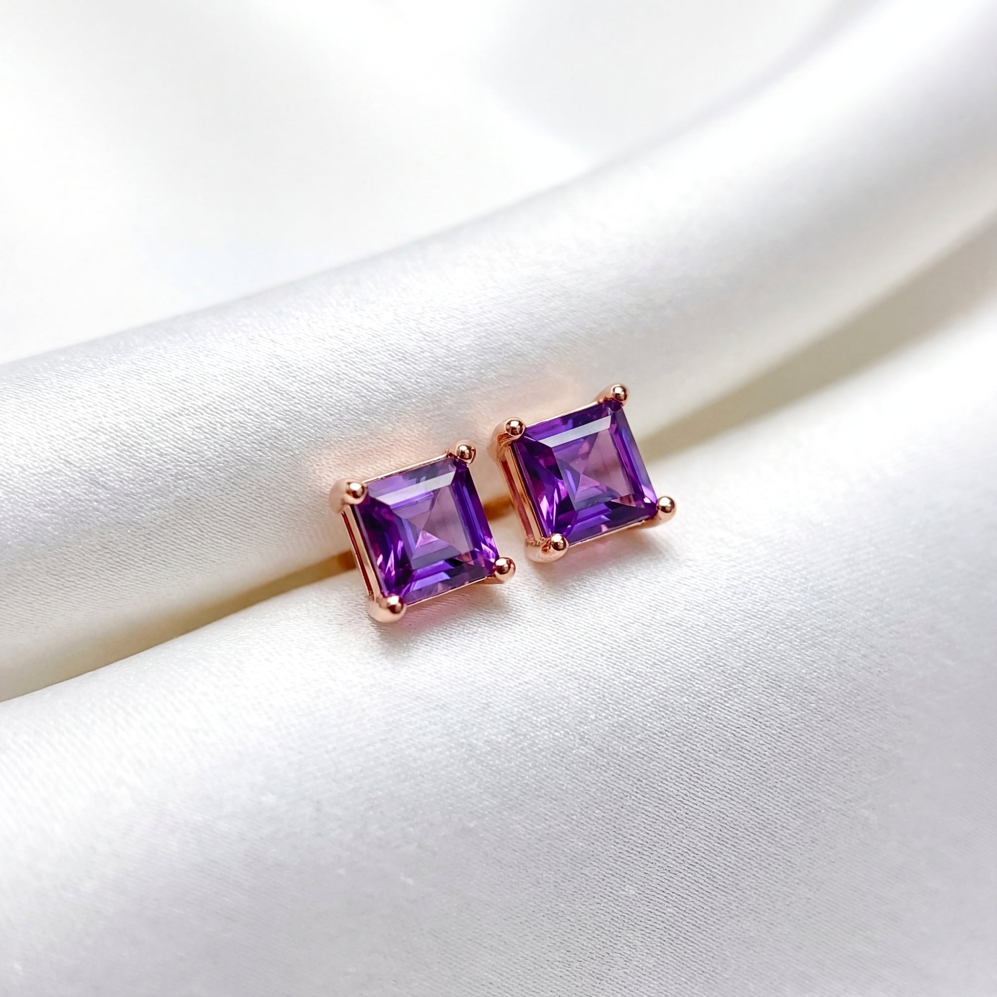 Square cut simulated amethyst stud earrings in 18K rose gold vermeil resting on white silk fabric.