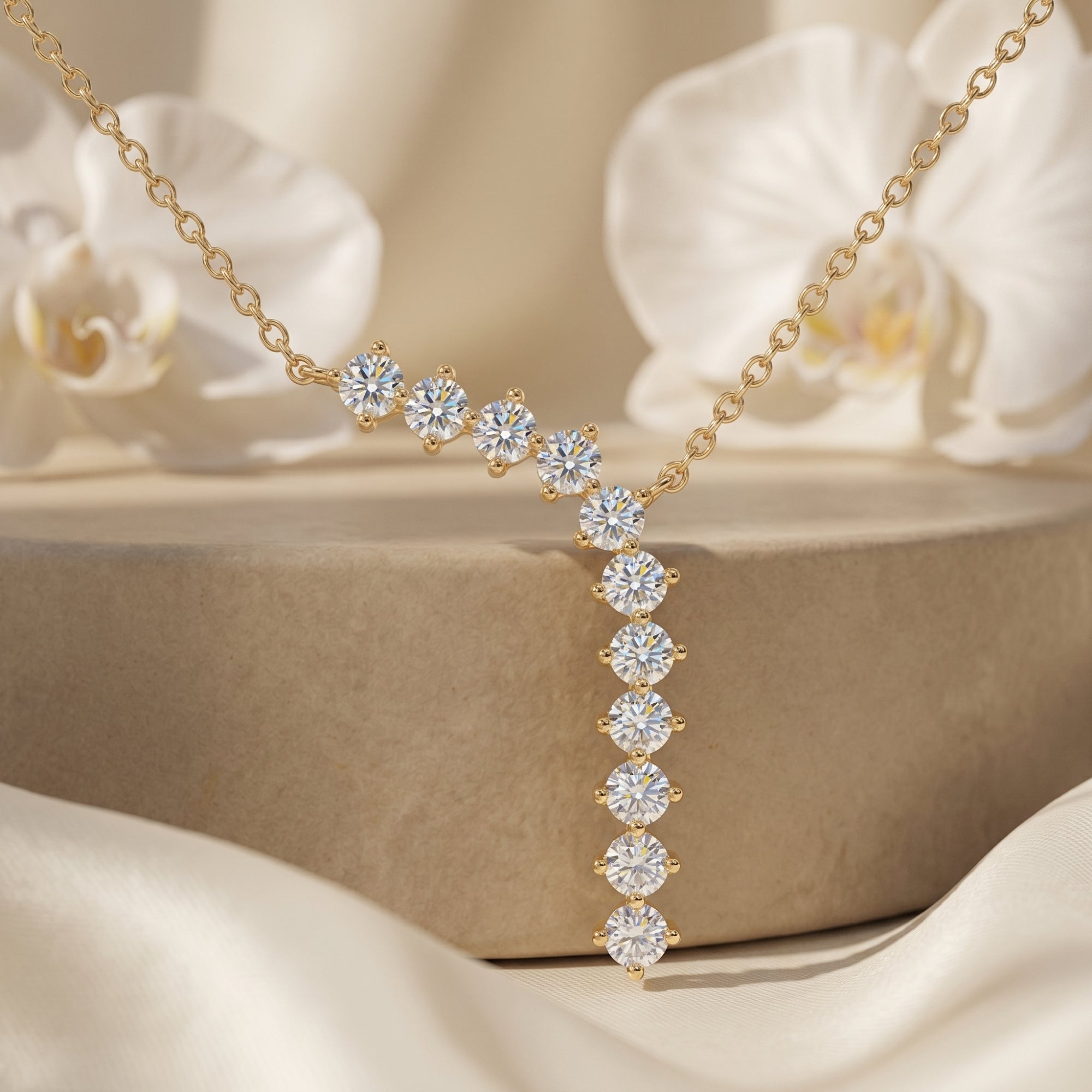 Gold curved drop necklace with round-cut moissanite stones on a stone pedestal with white orchids.