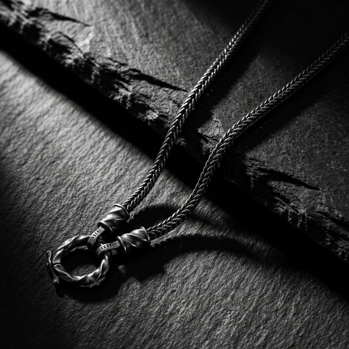 Silver twisted ring necklace and braided chain resting on dark slate stone.