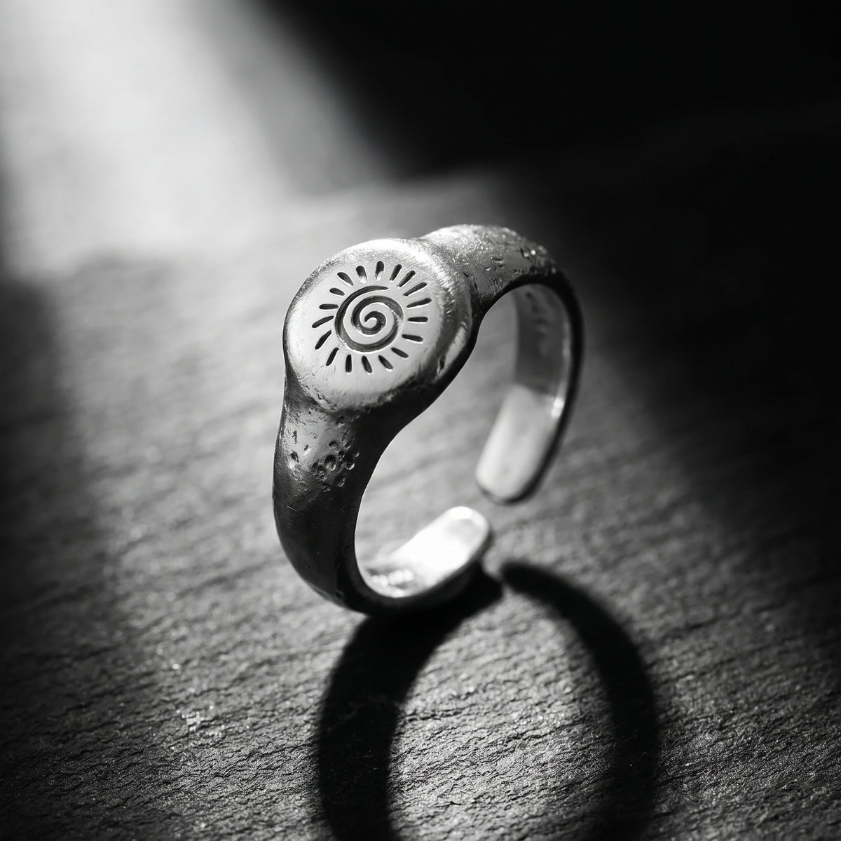 Silver solar spiral signet ring standing upright on a dark textured stone surface with a cast shadow.