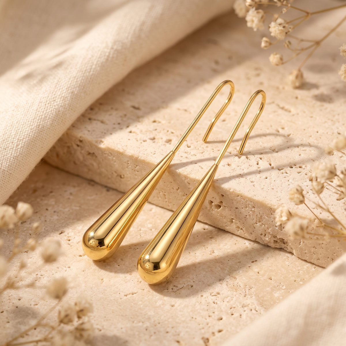 Gold-toned long teardrop earrings resting on textured stone blocks beside soft fabric.