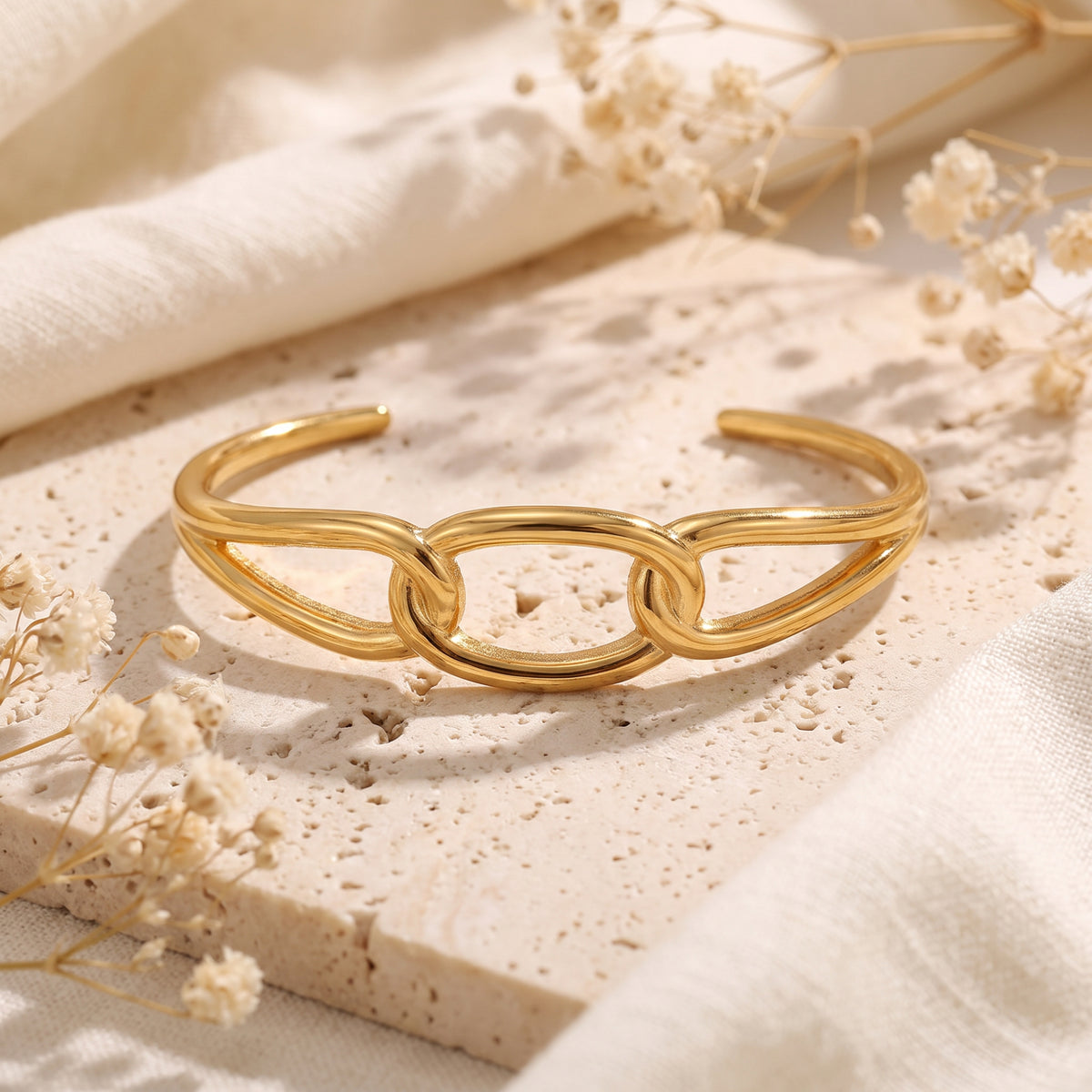 Gold-tone open cuff bracelet with linked oval loops displayed on a porous stone slab with dried flowers.