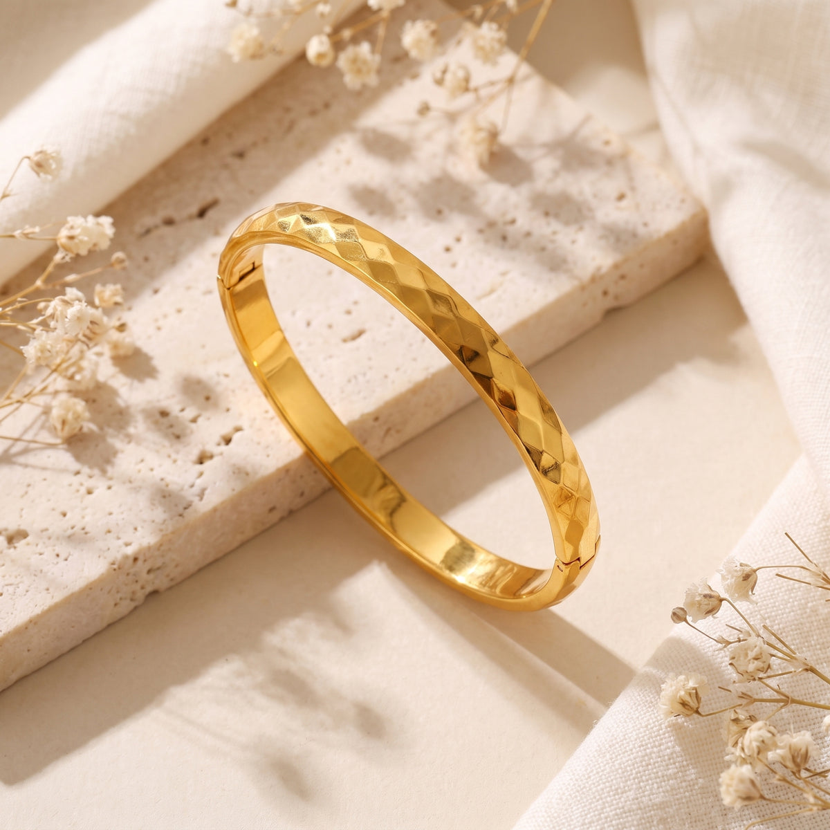 Gold-toned faceted bangle bracelet displayed at an angle on a light stone block with dried flowers.