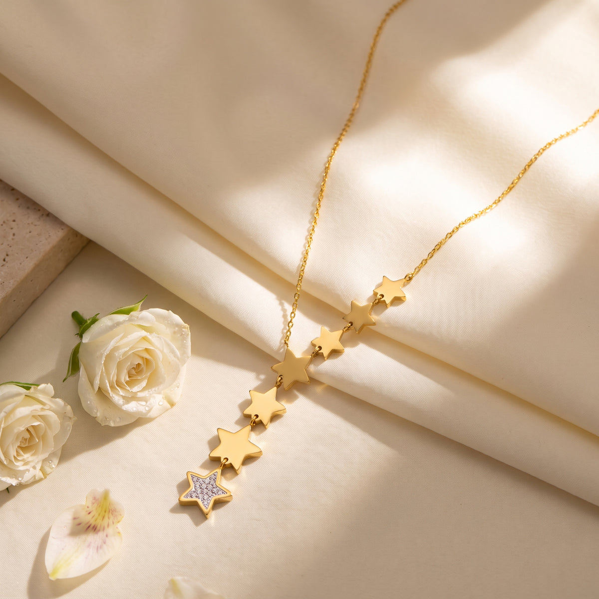 Gold-tone cascading star lariat necklace with a stone-paved star tip on cream fabric beside white roses.