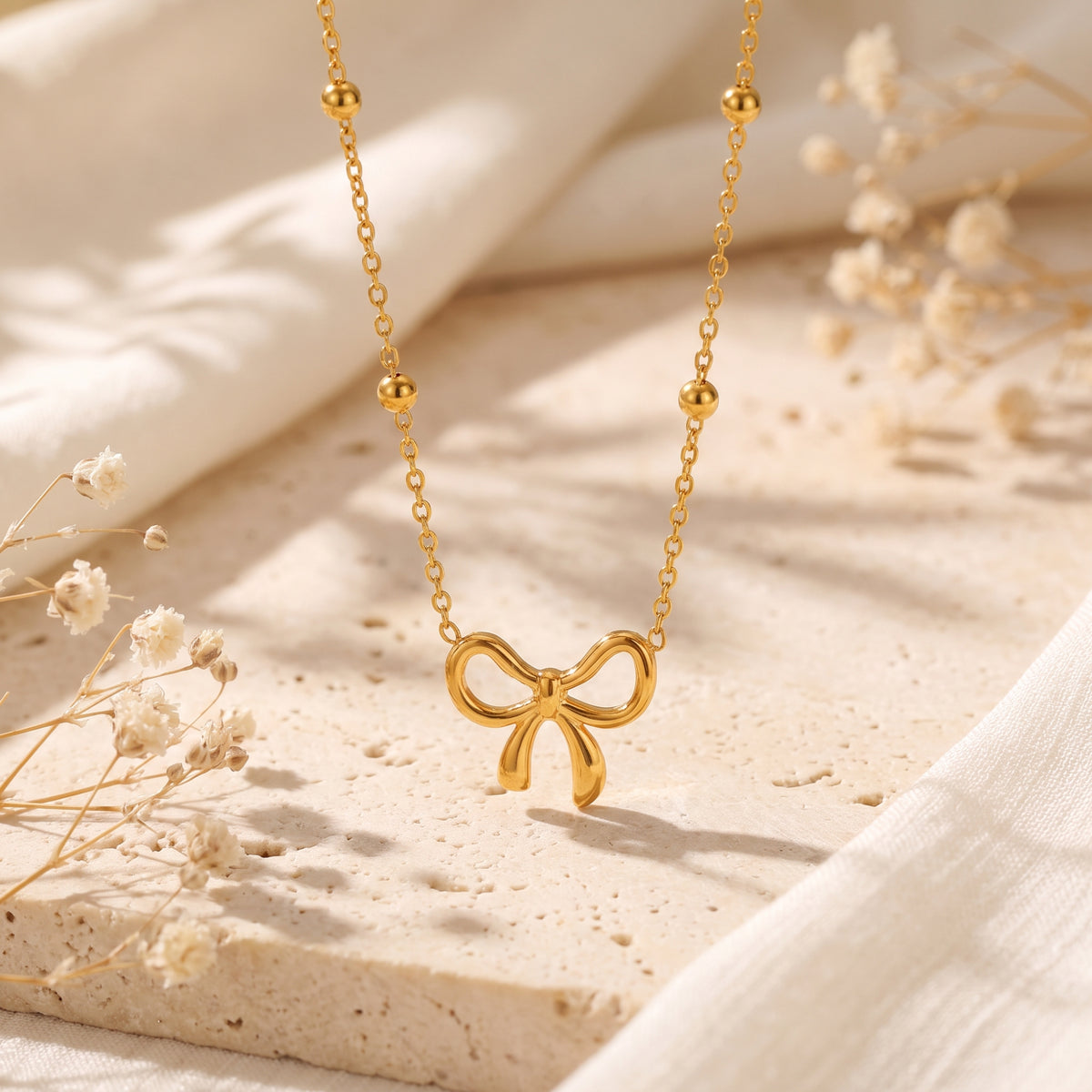 Gold-tone bow pendant on a beaded station chain, suspended above a beige textured surface with dried flowers.