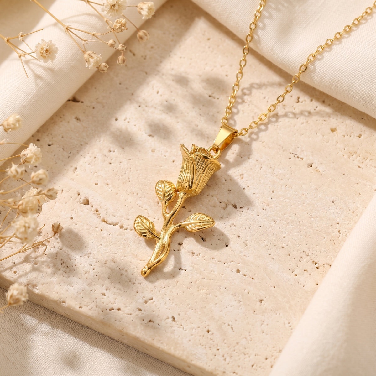 Detailed gold-tone rose pendant with stem and leaves lying flat on a textured stone surface.