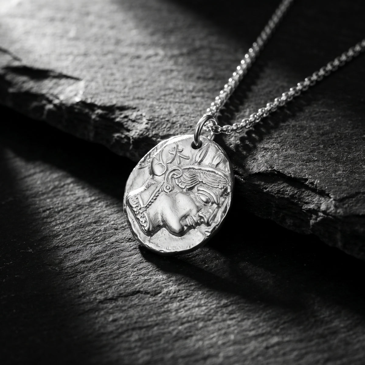 Close-up of silver Athenian coin pendant with a profile relief resting flat on dark slate stone.