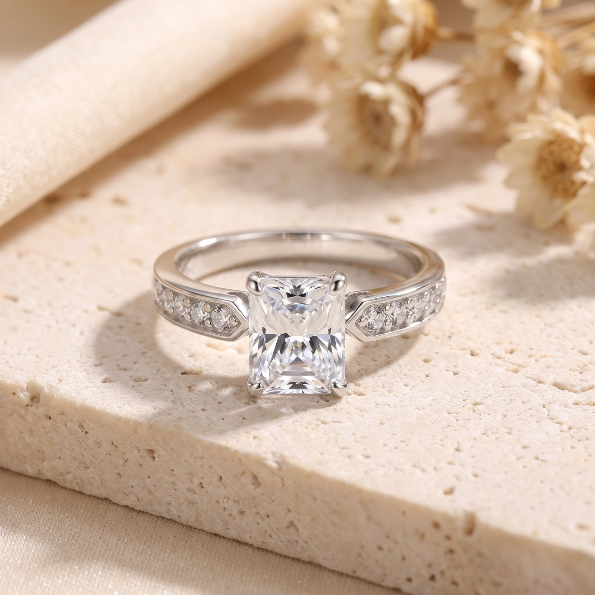Radiant-cut simulated diamond ring in S925 sterling silver on a stone surface with dried flowers.