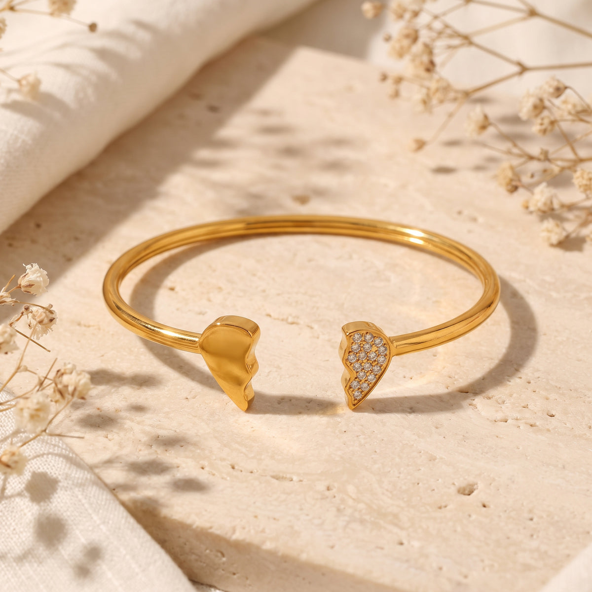 Gold-tone open cuff bracelet with two broken-heart ends, one smooth and one pavé set with crystals, on a stone slab.