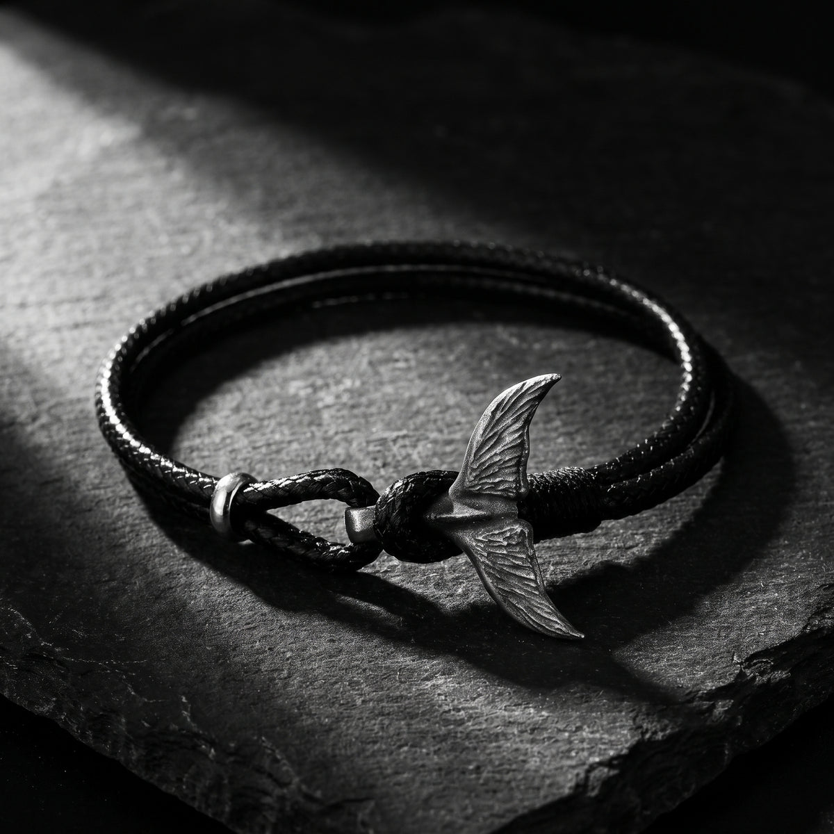 Close-up of a black cord bracelet with a silver whale tail clasp resting on a dark slate surface.