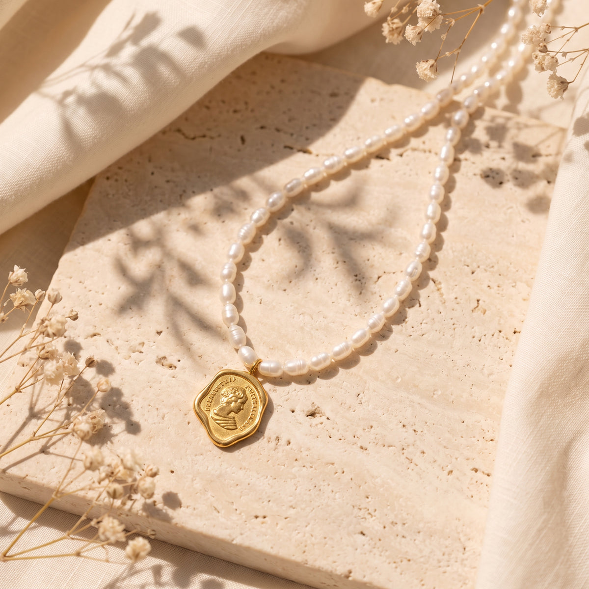 Gold-tone coin medallion necklace with white pearl-inspired beads on a textured stone surface with dried flowers.