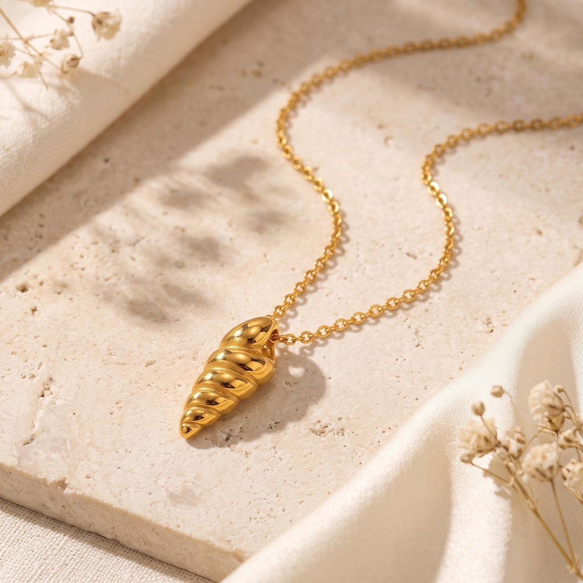 Gold-tone spiral shell pendant necklace displayed on travertine stone with cream fabric and dried flowers.