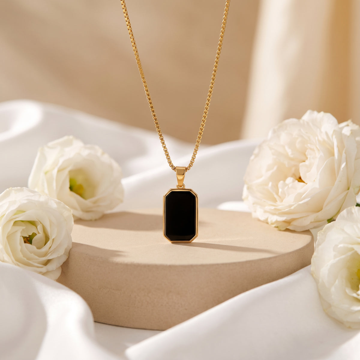 Gold-tone box chain necklace with an octagonal black pendant displayed upright on a beige pedestal among white flowers.