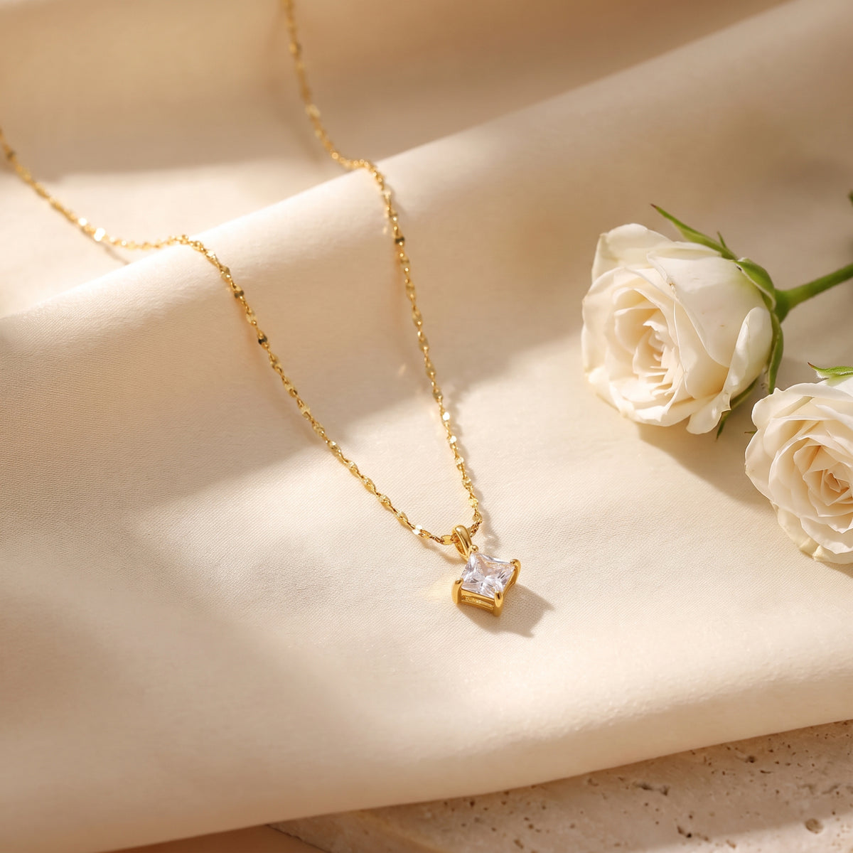 Gold-tone square solitaire pendant necklace draped over cream fabric beside two white roses.