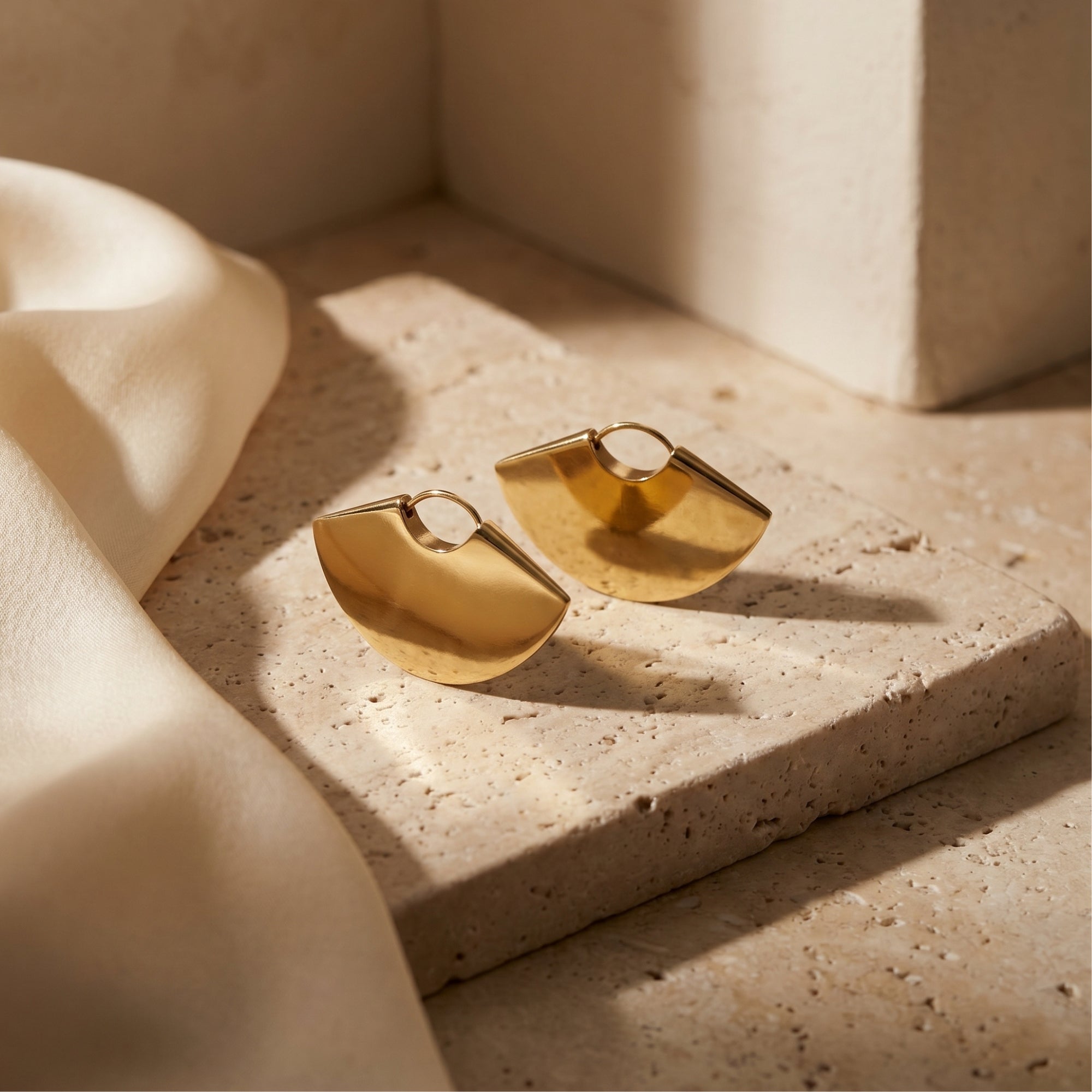 Gold fan earrings in 18K gold PVD stainless steel on a stone surface with silk.