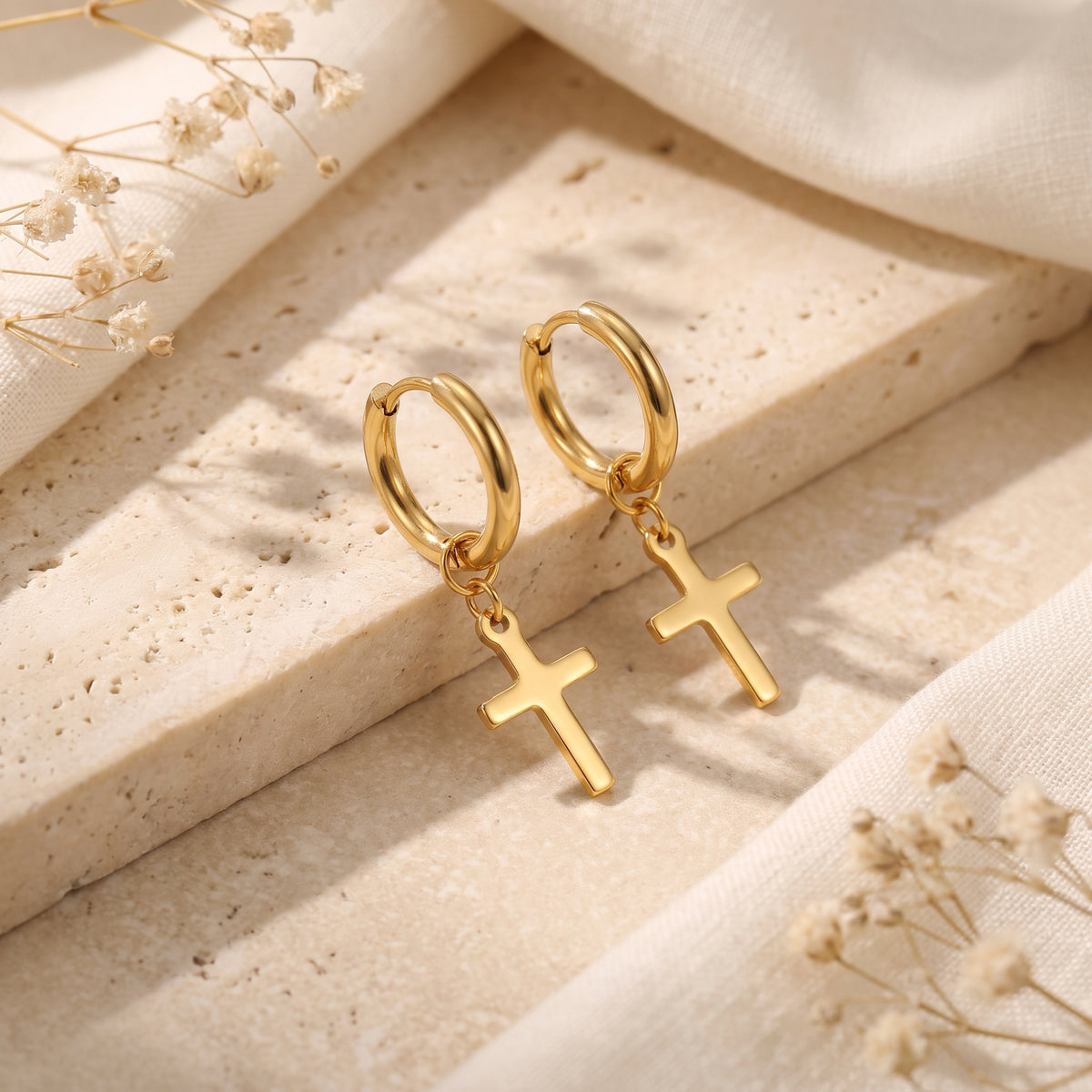 Pair of gold-tone huggie hoop earrings with dangling cross charms displayed upright on a textured stone slab.