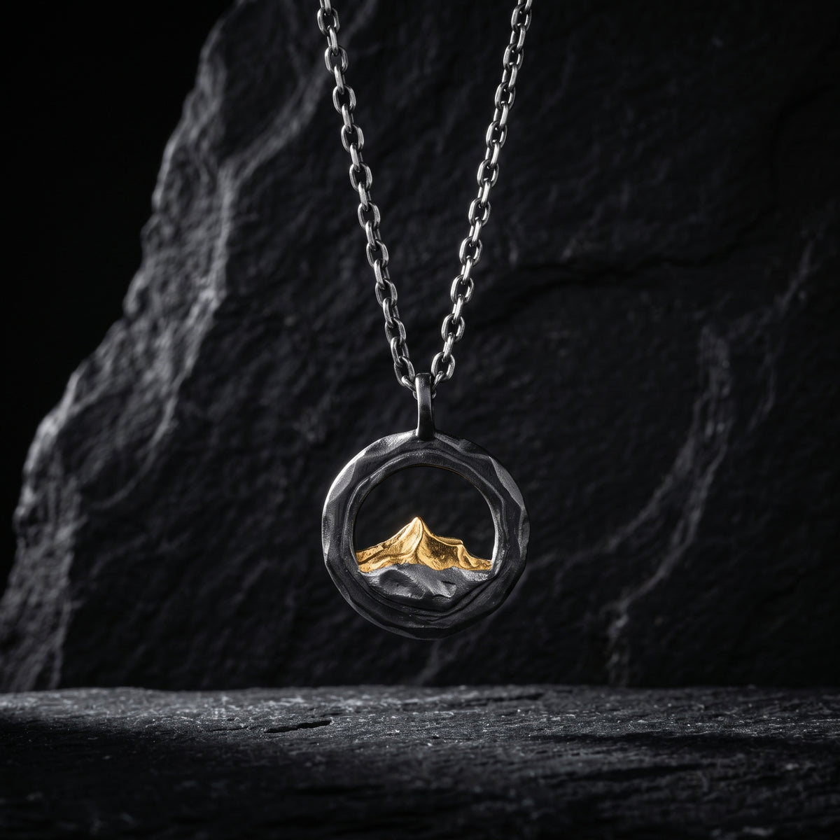 Front view of a circular silver pendant with a gold-tone mountain peak hanging from a chain against dark rock.