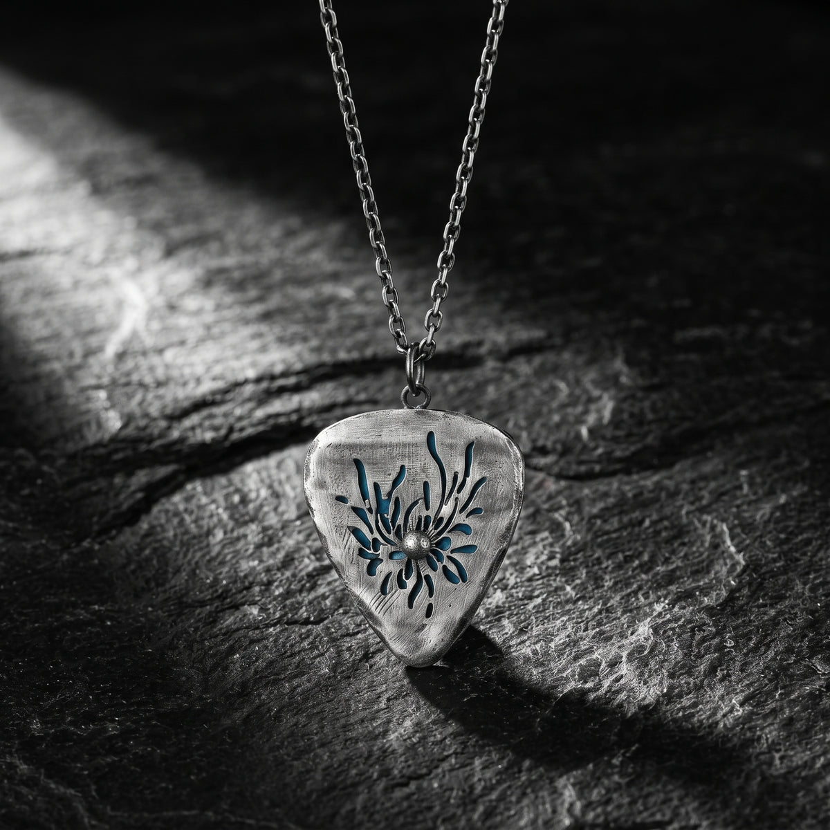 Silver guitar-pick pendant necklace with a carved anemone motif over a cyan backdrop on dark slate.
