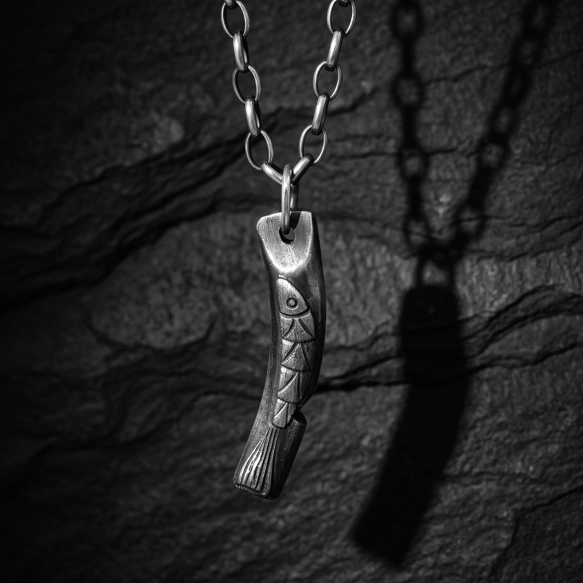 Silver carved fish bar pendant hanging vertically against a dark slate background.