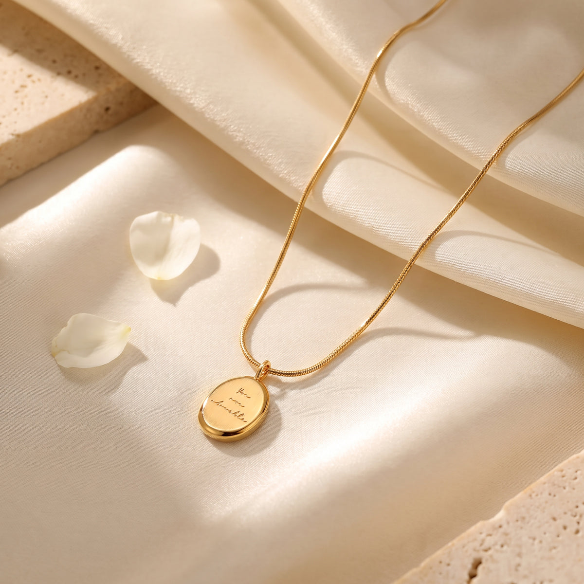 Gold-tone oval medallion necklace engraved with “You are lovable” on draped silk beside white rose petals.