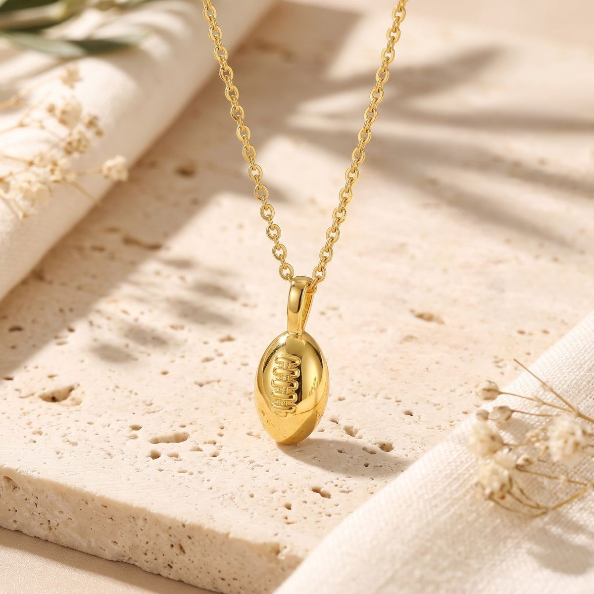 Close-up of a gold-tone football pendant necklace with raised laces resting on a light porous stone slab.