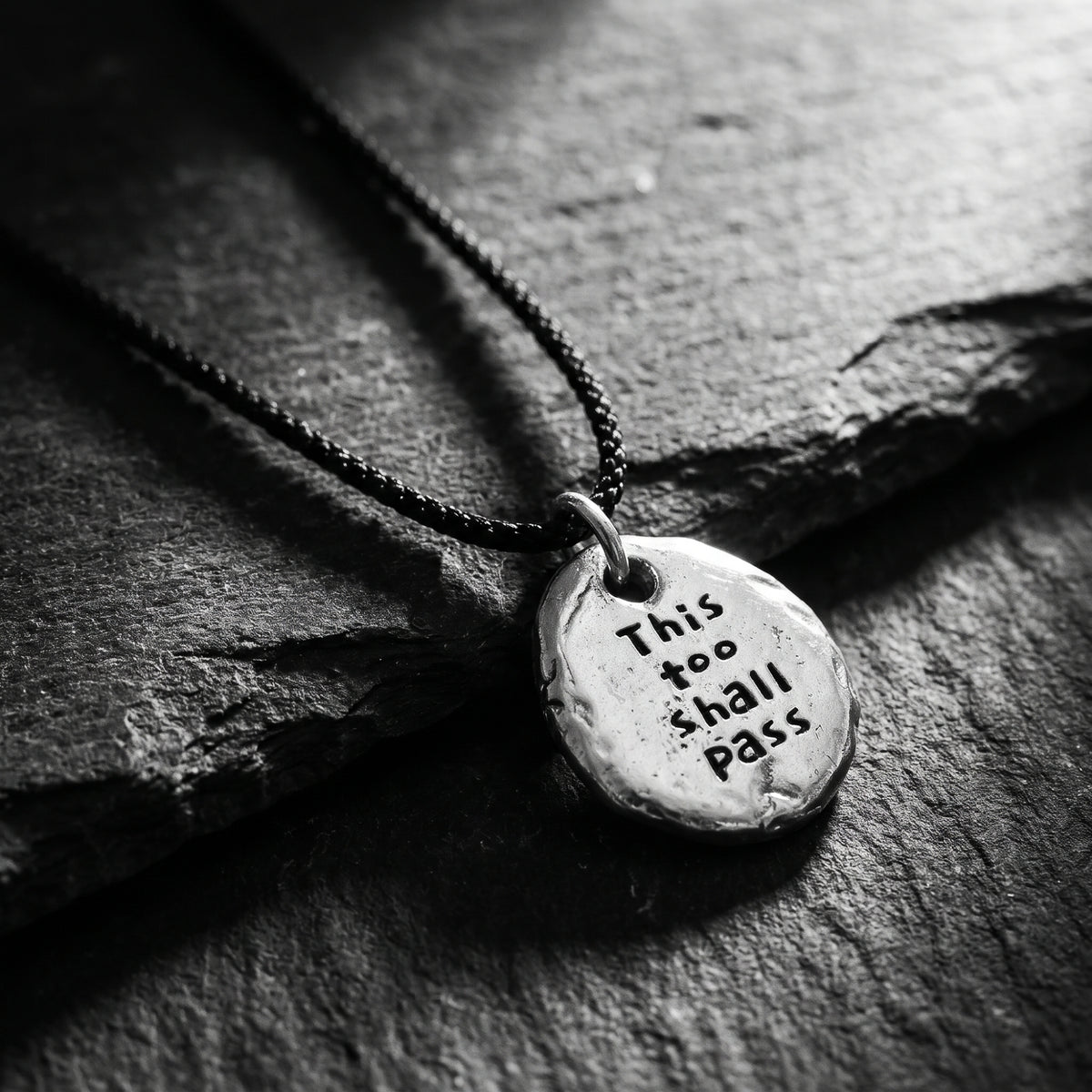 Close-up of a silver pebble pendant engraved “This too shall pass” on dark slate, with a black cord.