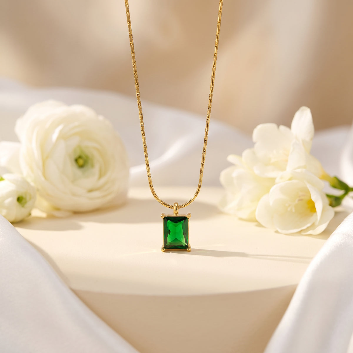 Gold-tone necklace with a rectangular emerald-green pendant displayed on a beige pedestal beside white flowers.