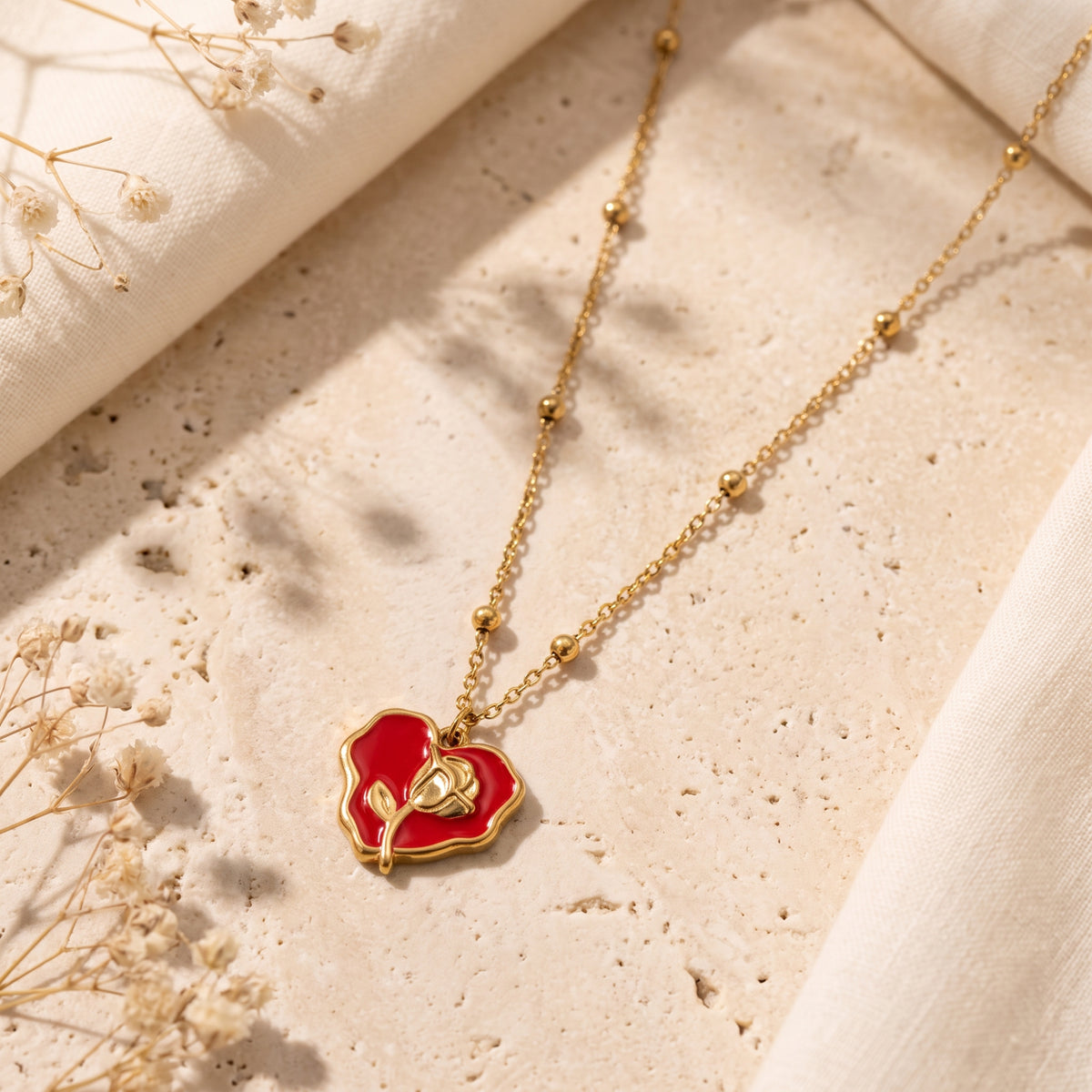 Flat lay of a gold-tone beaded chain necklace with a red enamel rose heart pendant on a textured stone surface.