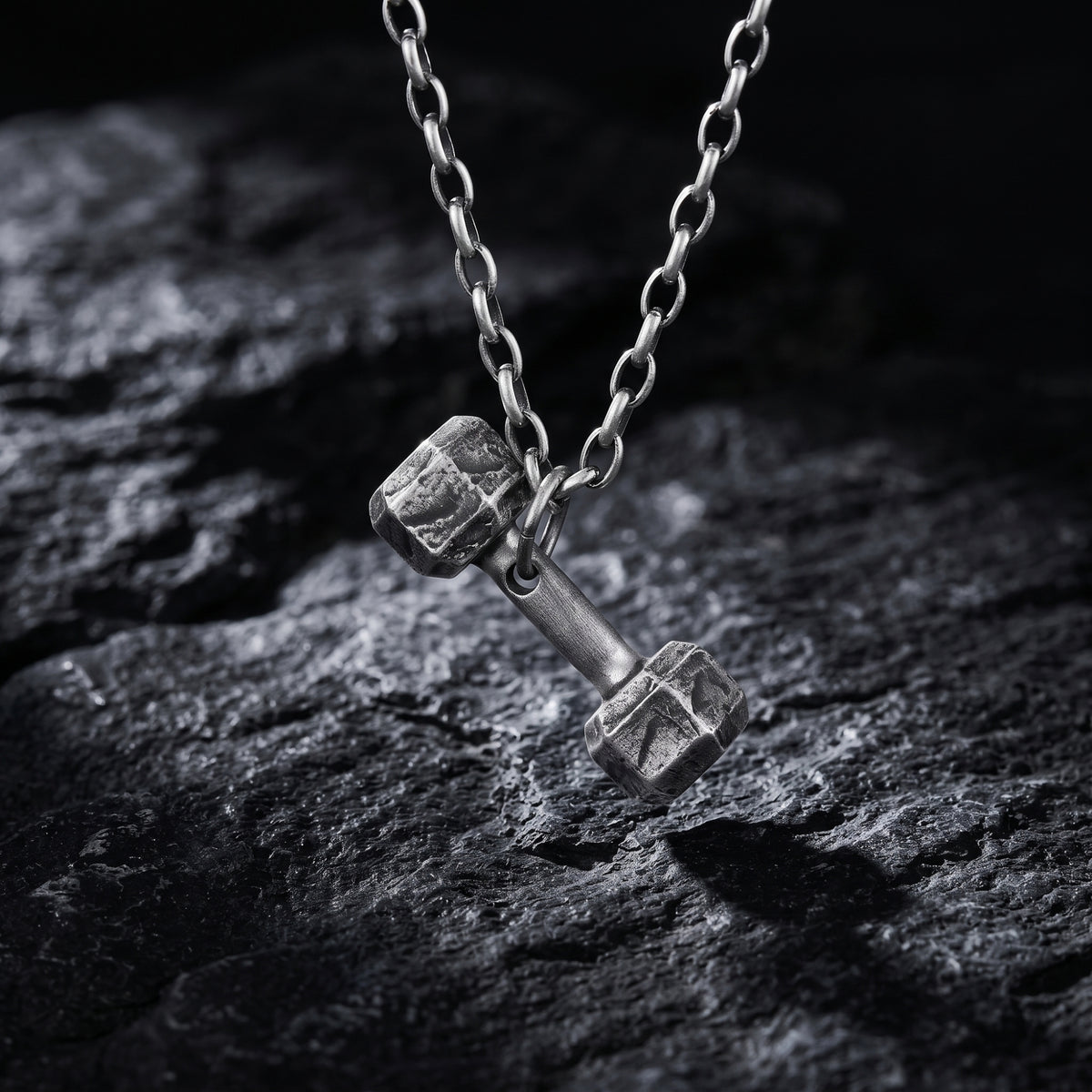 Close-up of a fine silver dumbbell pendant hanging from a chain over dark textured rock.