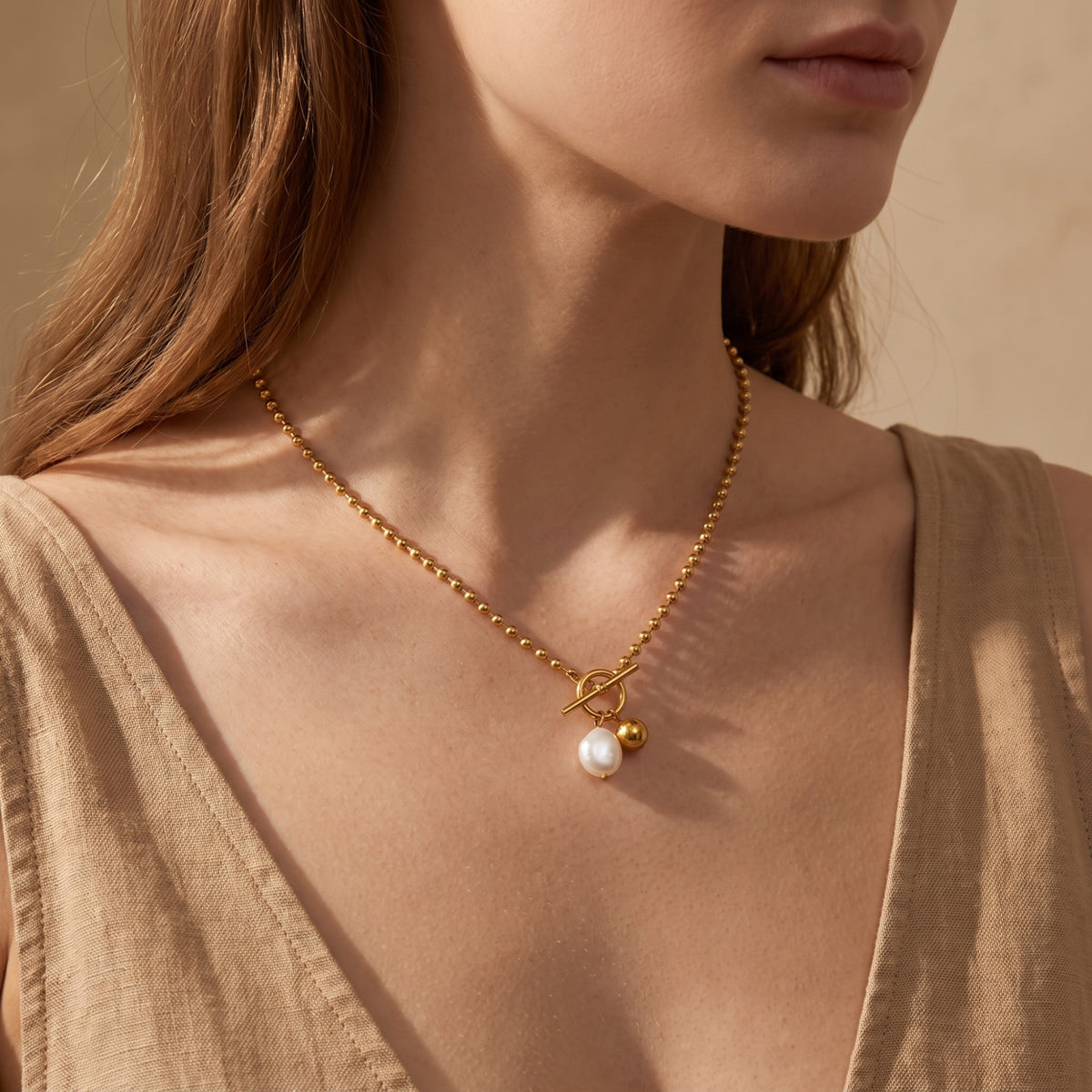 Model wearing a gold-tone ball chain toggle necklace with pearl and sphere charms over a tan top.