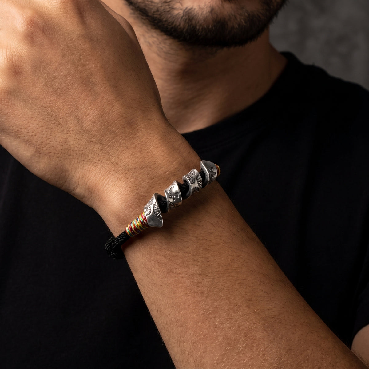 Close-up of a person wearing a black cord bracelet with multiple engraved silver sections and colorful thread accents.