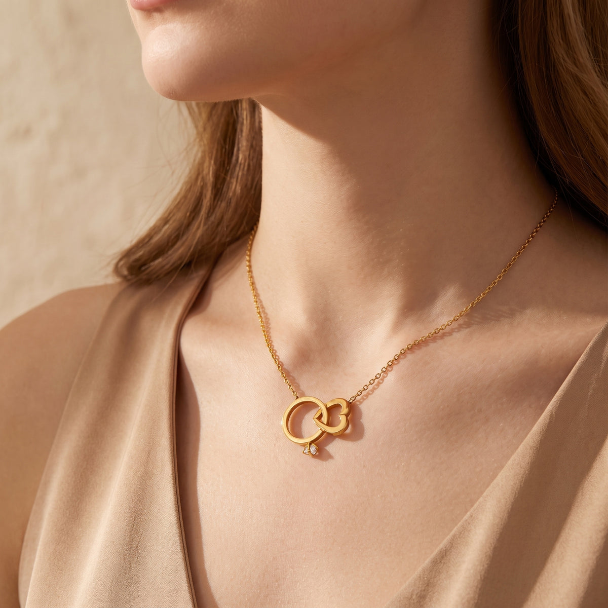 Model wearing the gold-tone interlocking ring and heart necklace over a tan blouse.