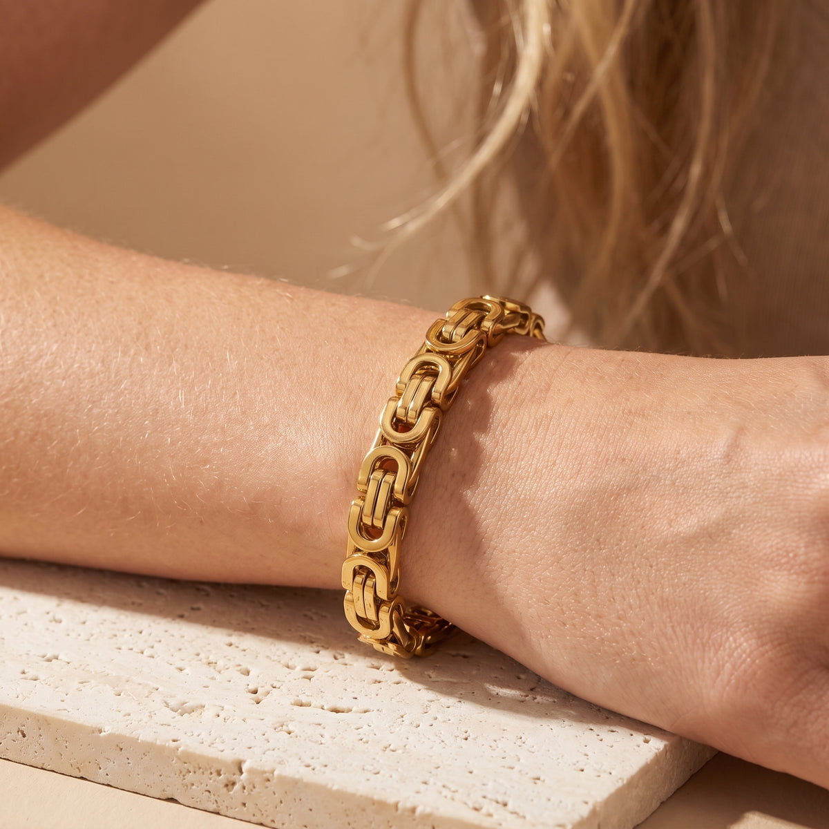 Close-up of the gold-toned Byzantine link bracelet worn around a wrist over a light stone surface.