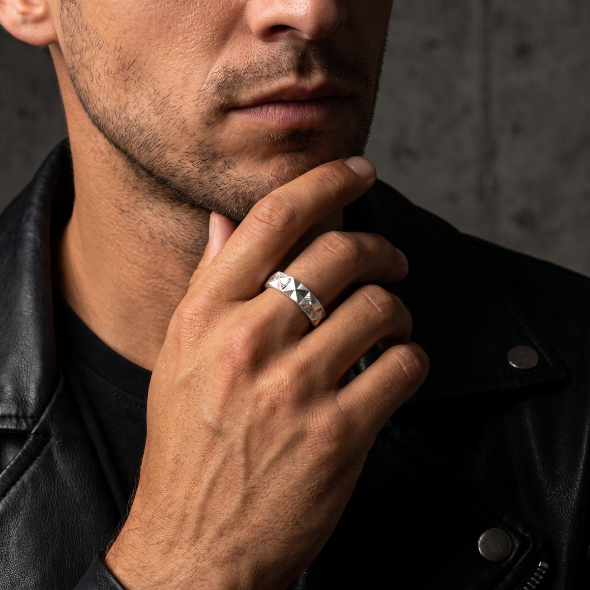Man wearing a silver faceted pyramid ring while touching his chin, dressed in a black leather jacket.