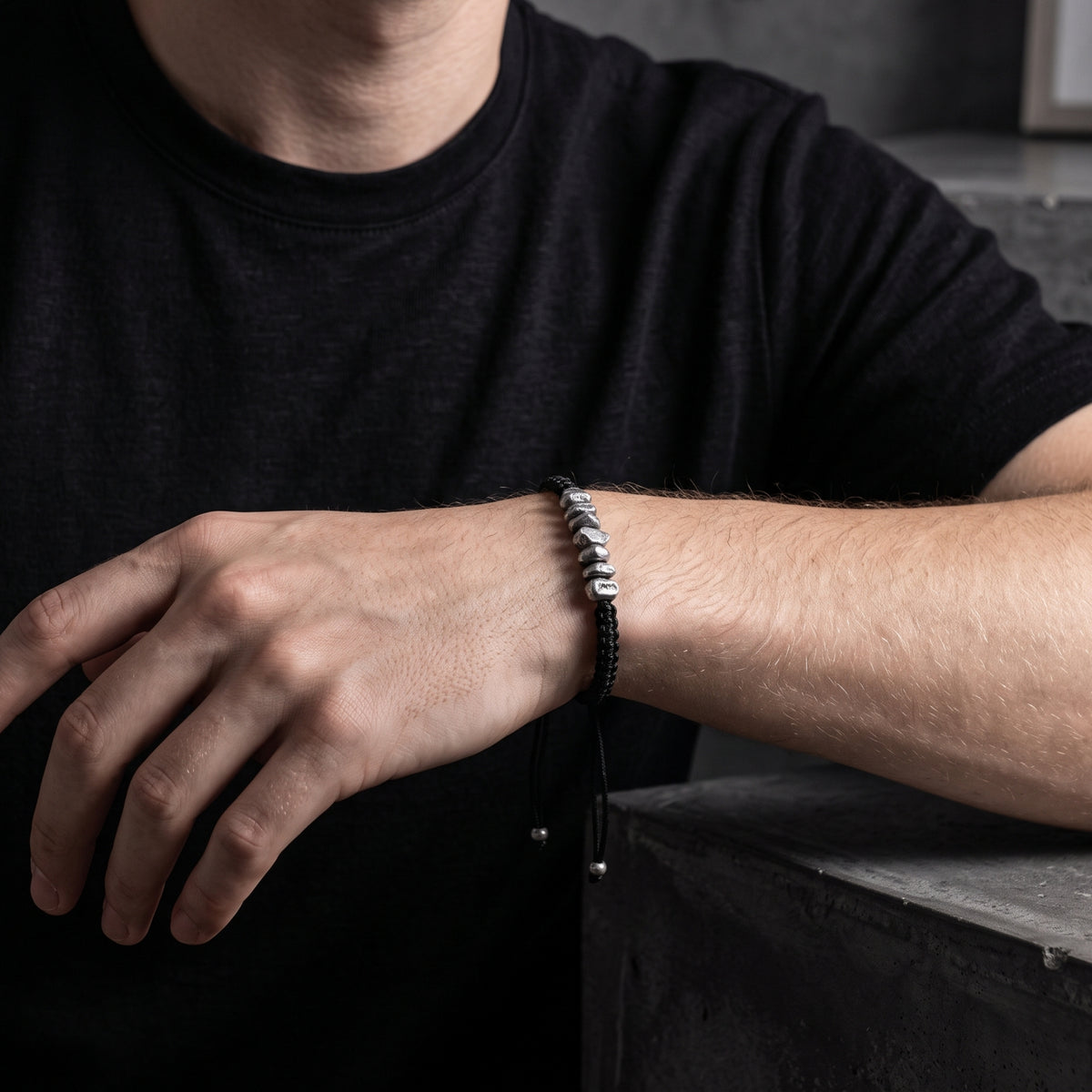 Black cord bracelet with silver nuggets worn on a man's wrist above a gray block.