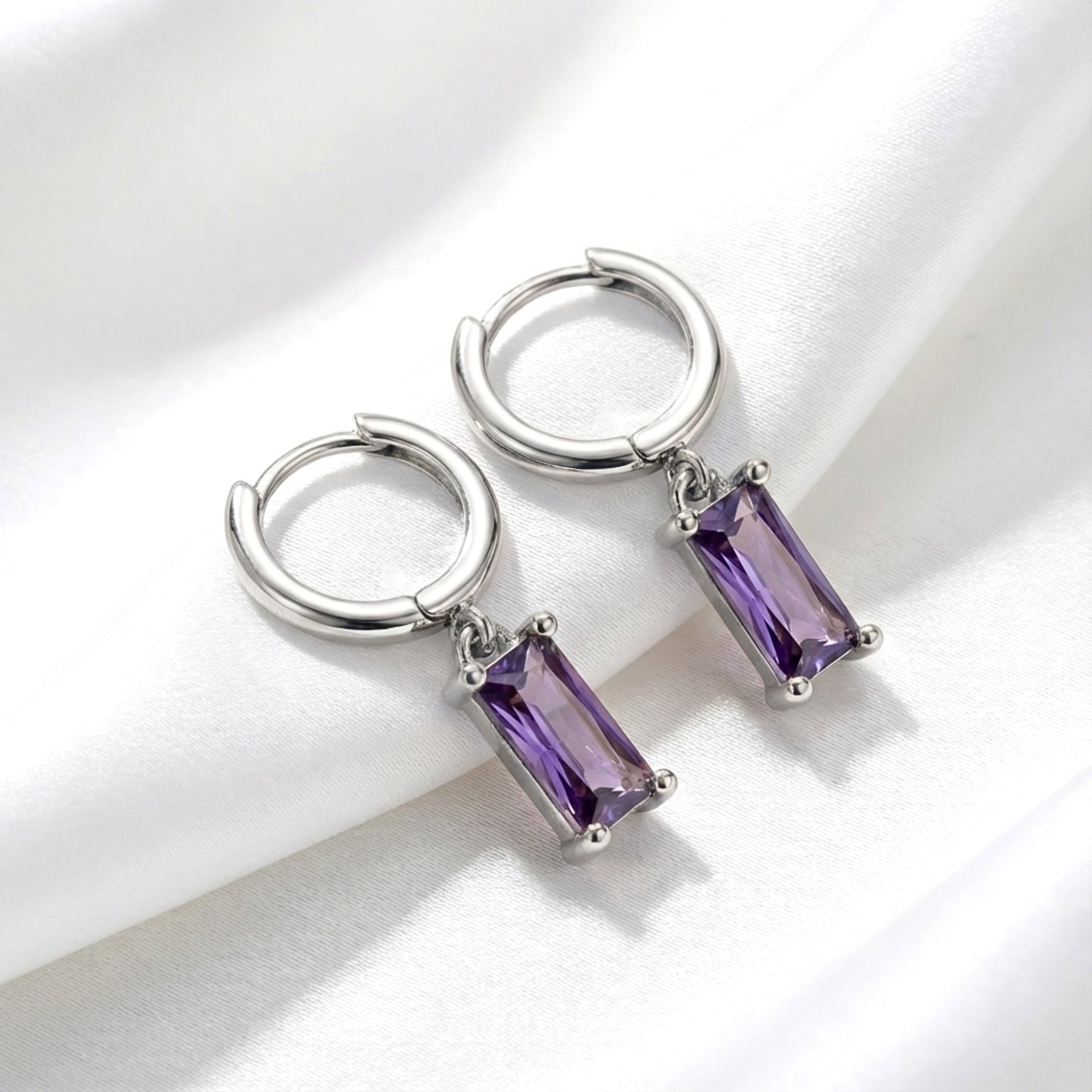 Sterling silver hoop earrings with emerald cut simulated amethyst drops resting on white fabric.