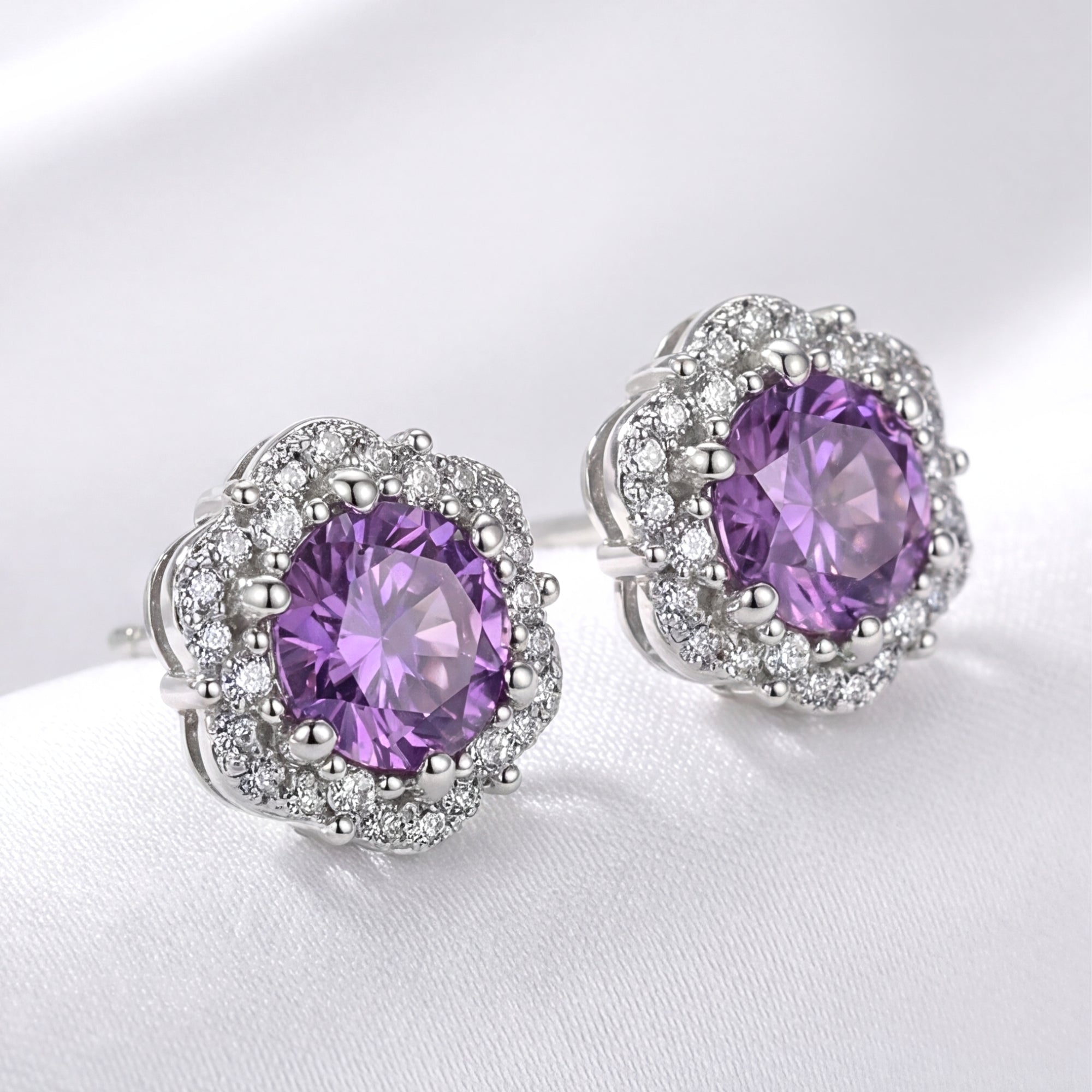 Angled view of round cut simulated amethyst floral halo stud earrings in S925 sterling silver on white fabric.