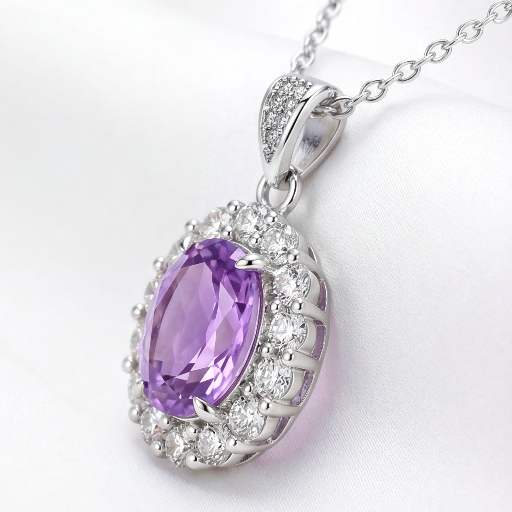 Side profile of an oval simulated amethyst halo pendant in sterling silver on white silk.