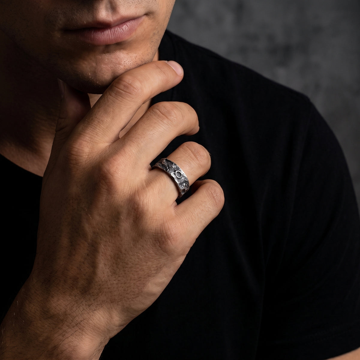 Textured silver open-band ring worn on the model’s ring finger as his hand rests against his chin.