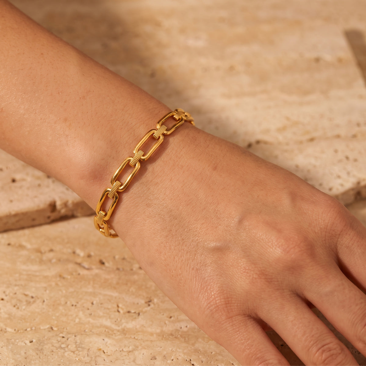 Gold-tone paperclip link bracelet worn on a wrist in close-up over a pale stone surface.