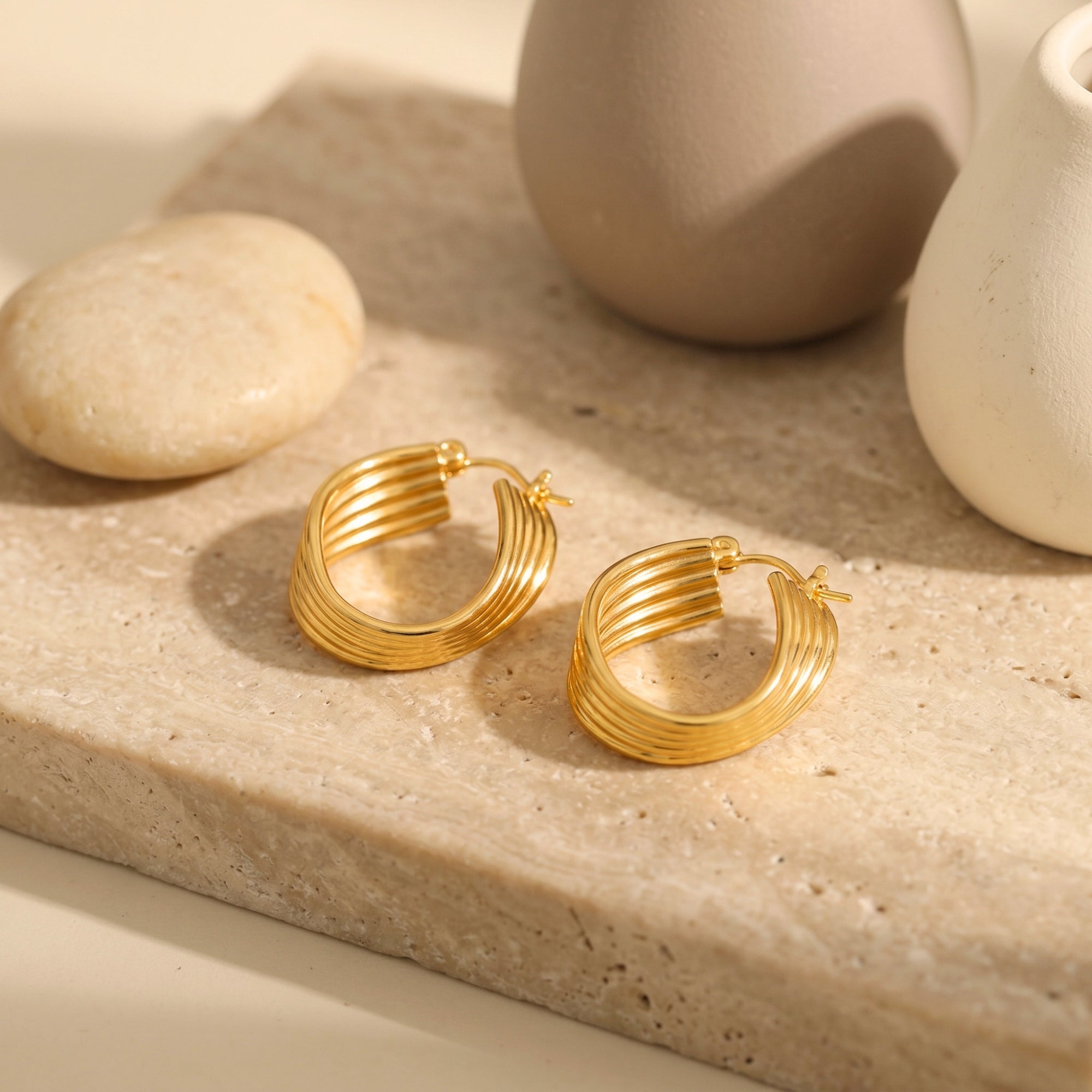 Gold layered architectural hoop earrings on a stone surface.