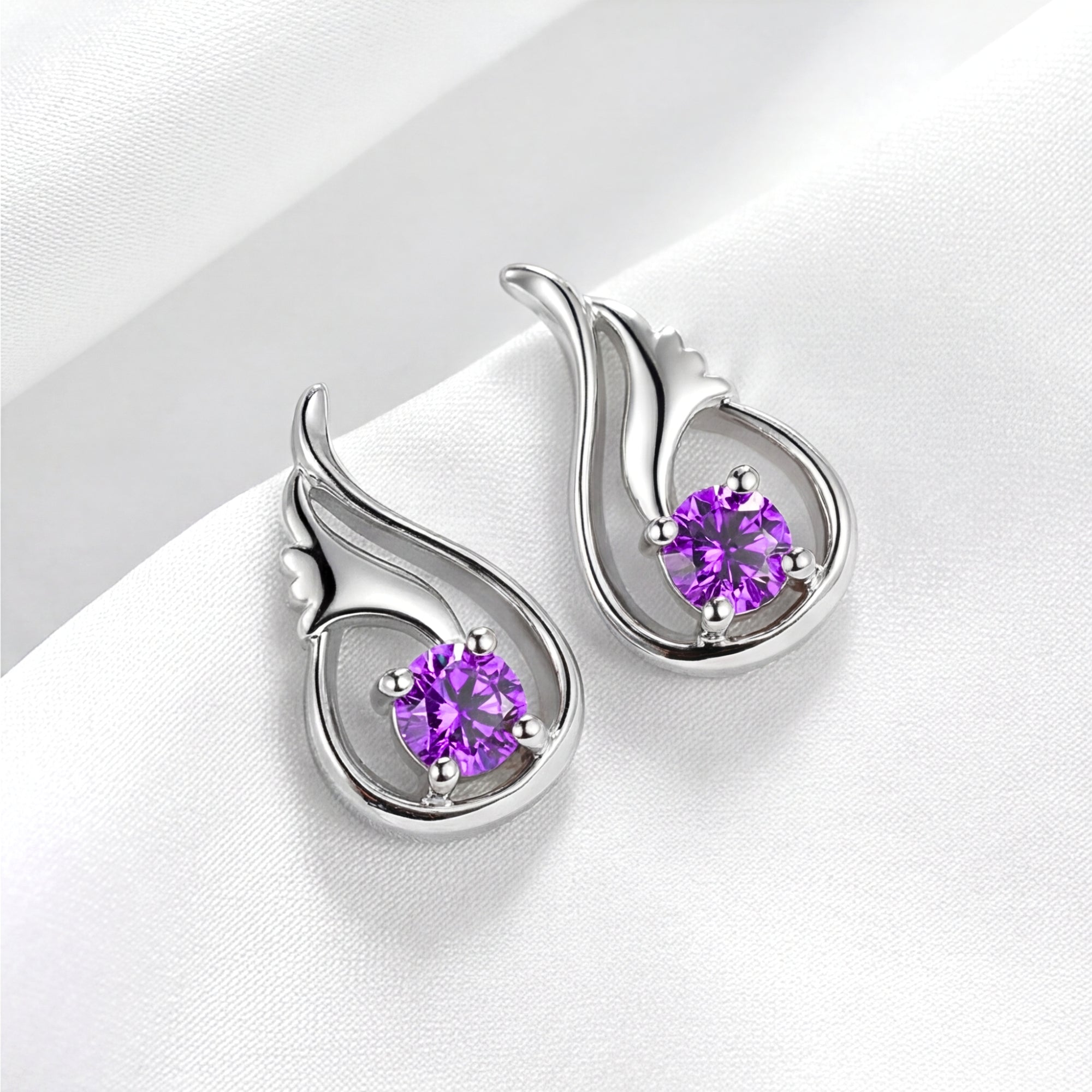 Angled view of sterling silver Lysithea drop earrings with round violet simulated amethyst on white fabric.