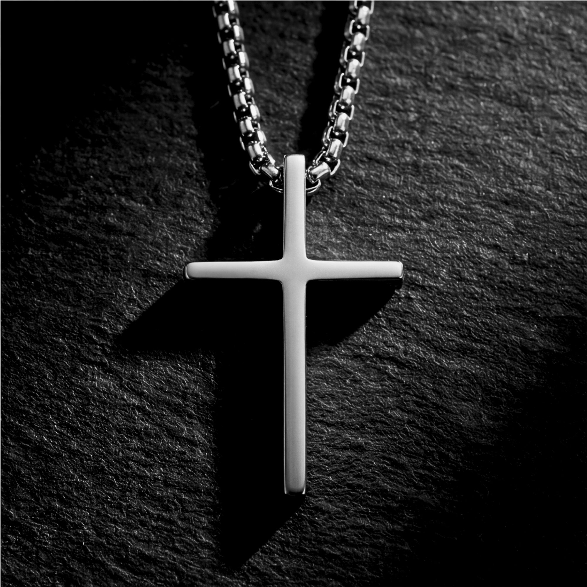 Close-up of the silver stainless steel Devotion Cross pendant on a box chain against a dark stone background.