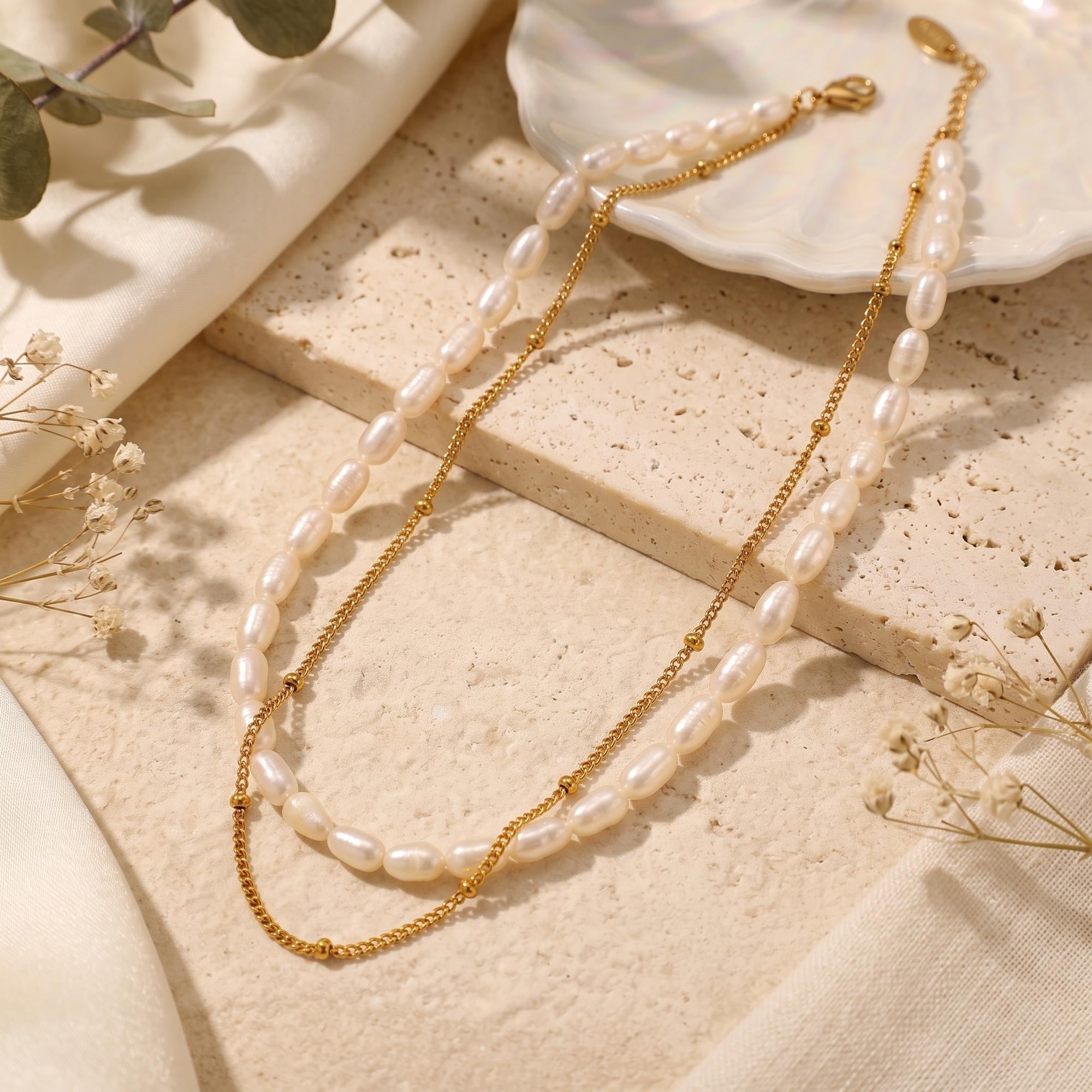 Gold layered necklace with freshwater pearls and satellite chain on a textured stone surface.