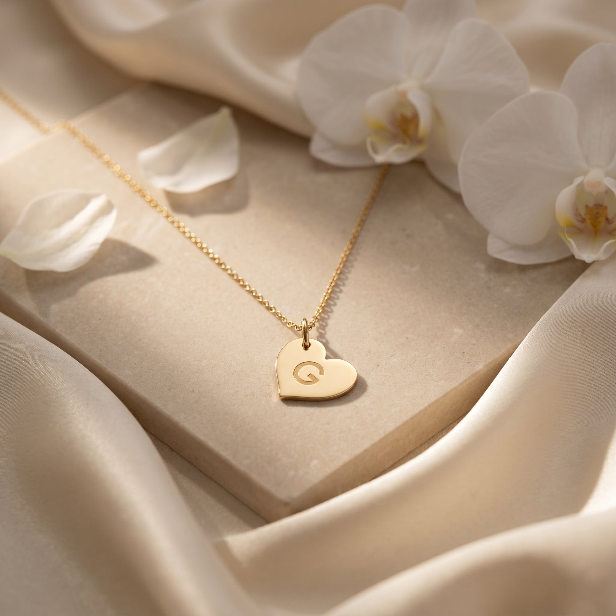18K gold heart pendant necklace with engraved 'G' styled on a stone surface with white petals.