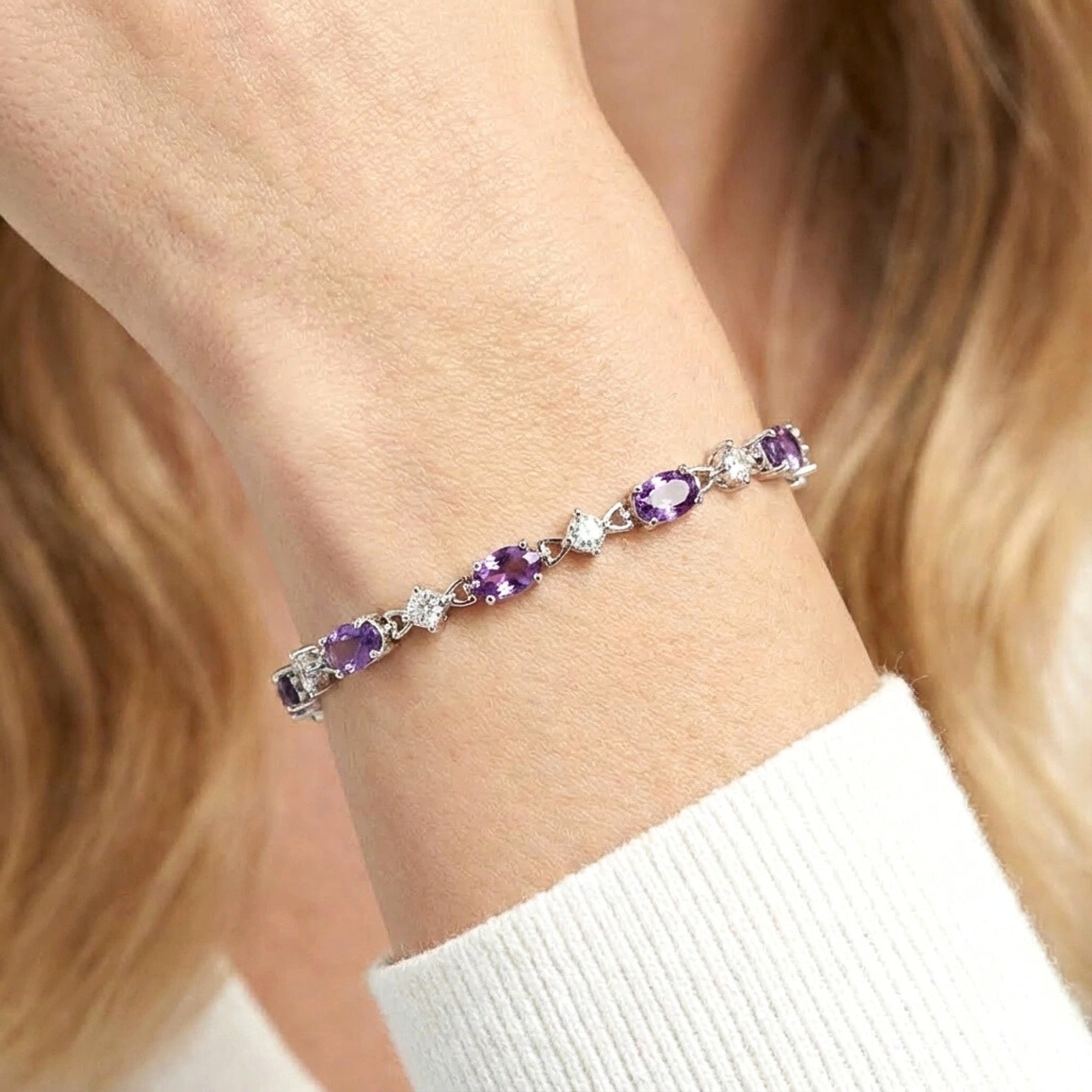 Close-up of oval cut simulated amethyst and round clear stones in a sterling silver tennis bracelet on a wrist.