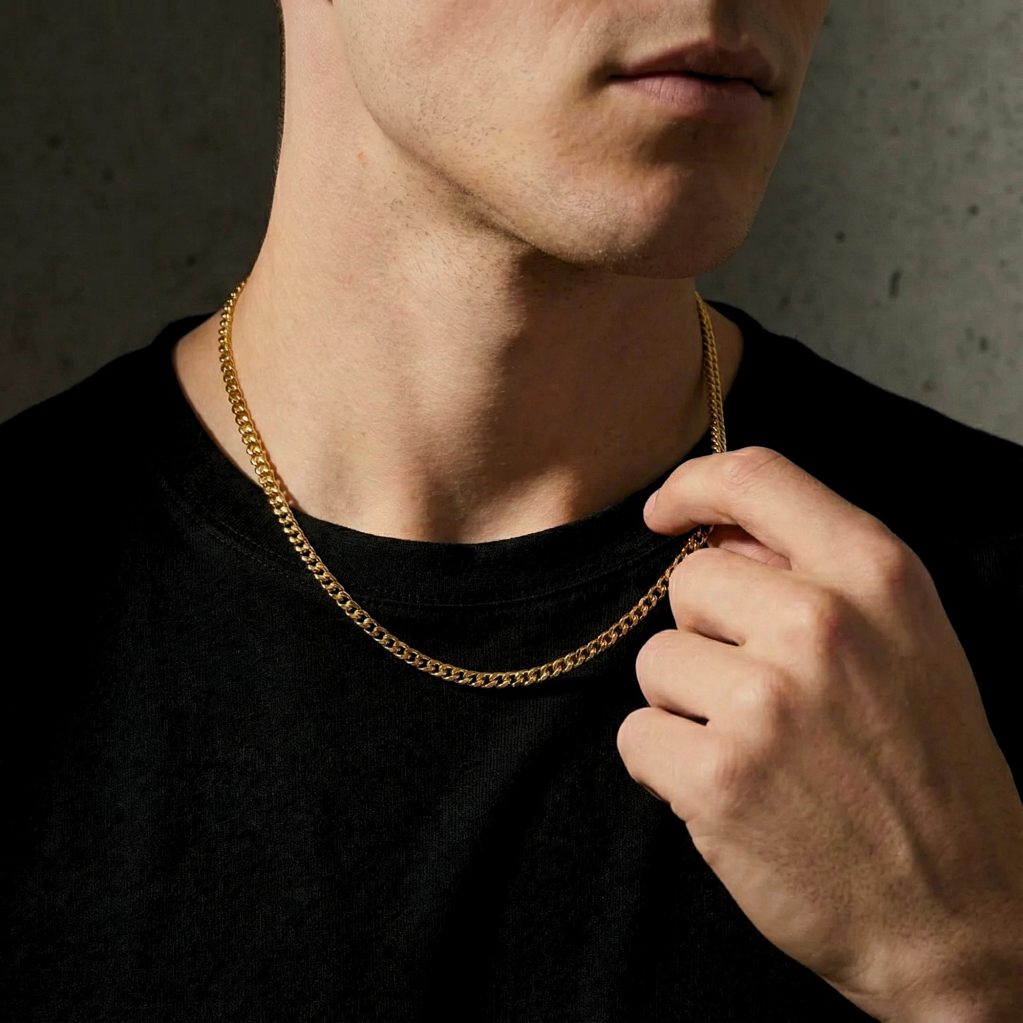 Close-up of a man wearing and touching a 4mm gold stainless steel Cuban chain necklace over a black t-shirt.
