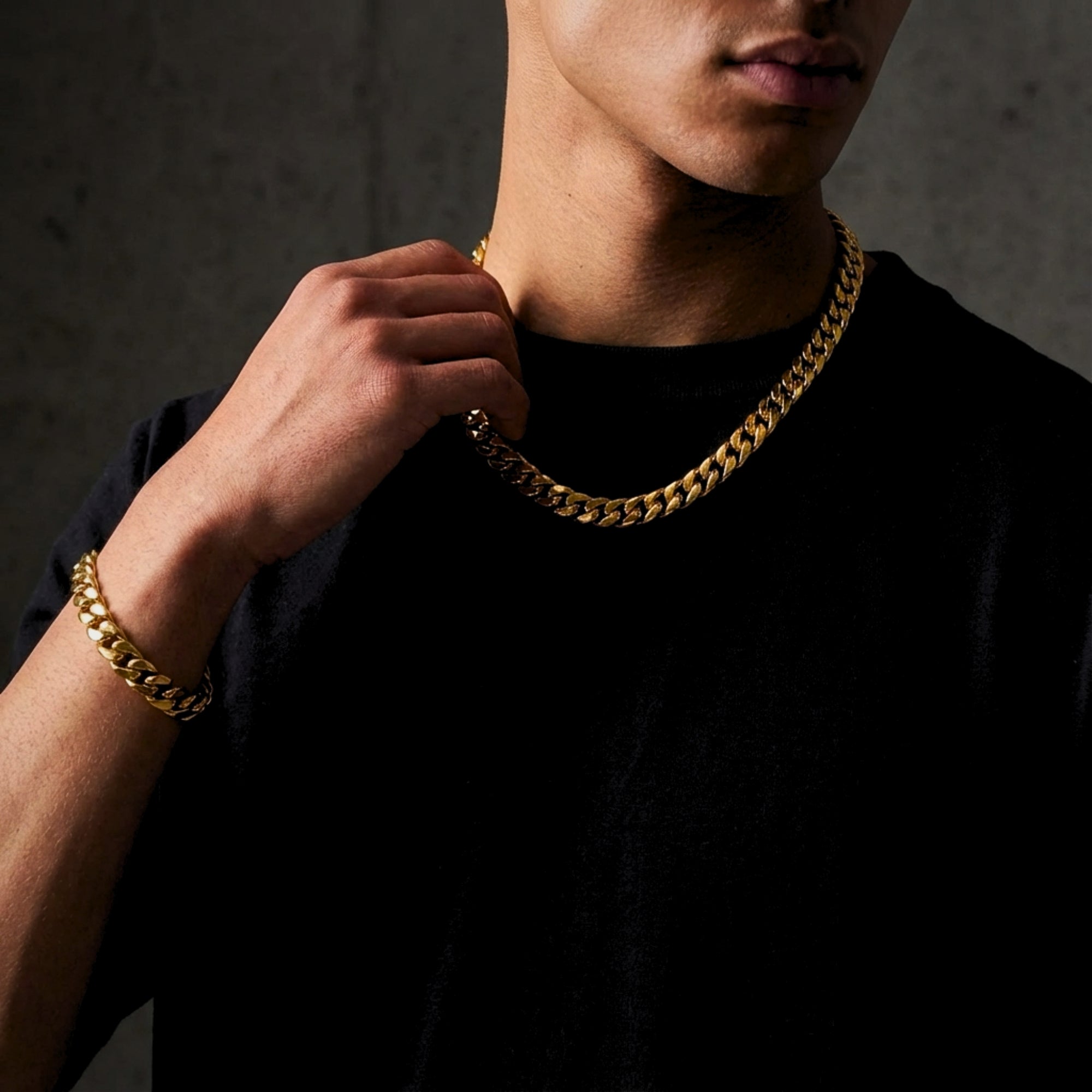 Model wearing the gold-tone stainless steel Cuban link chain and bracelet set against a concrete wall.