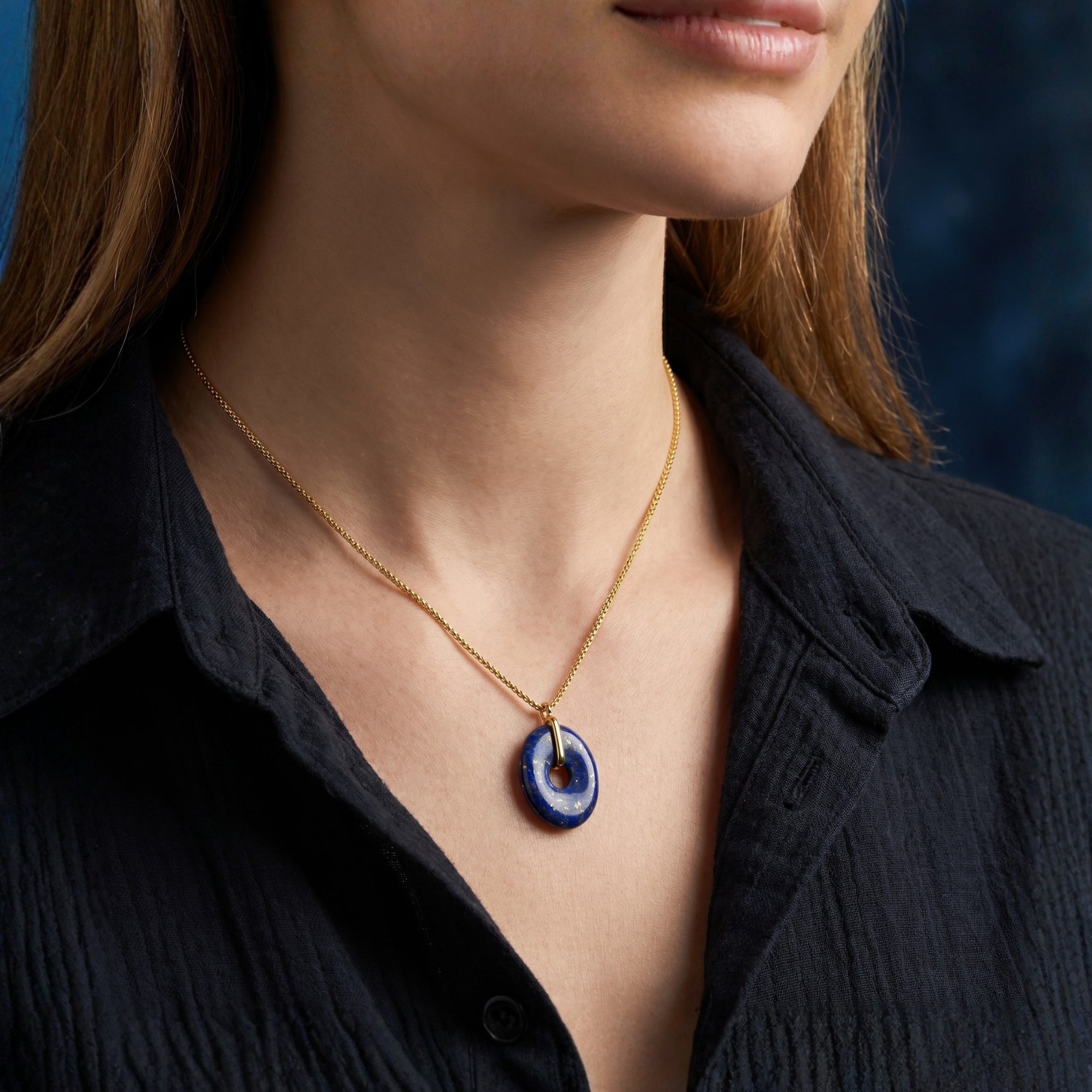 Natural lapis lazuli oval pendant on a gold plated chain worn by a model in a black button-down shirt.